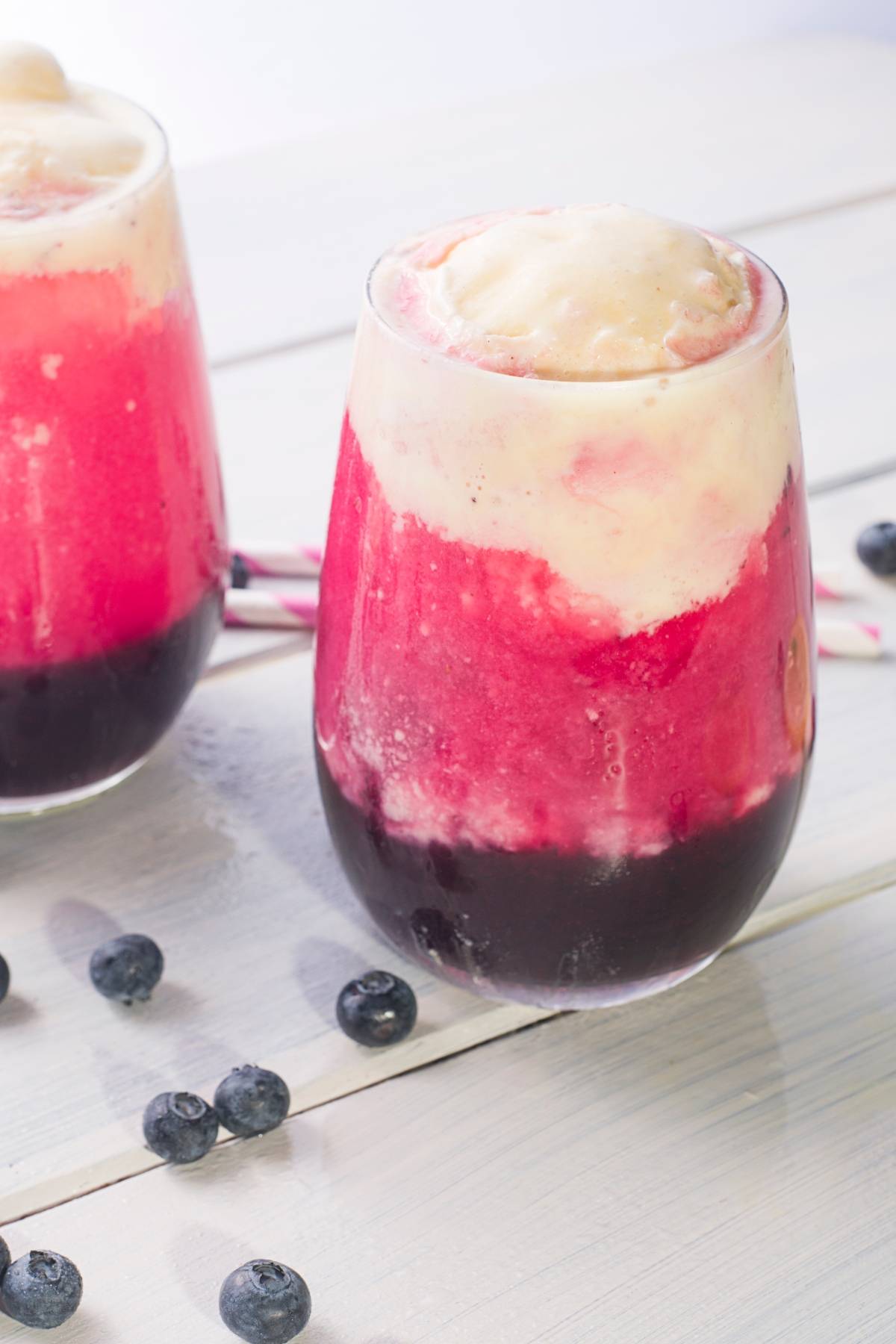 Two glasses of pink and purple sparkling blueberry float with striped straws and fresh blueberries sit on a white table, perfect for trying a new ice cream float recipe.
