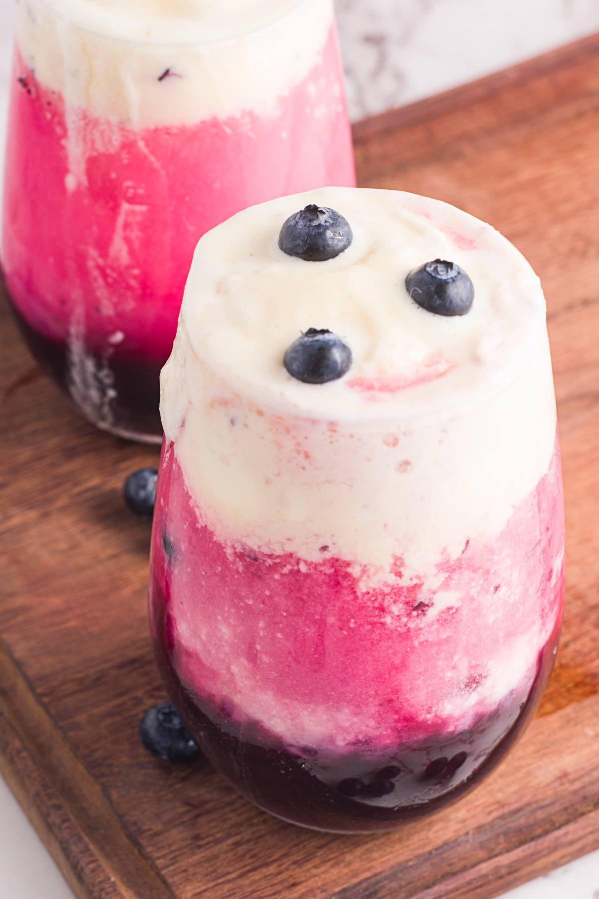 Layered pink and white smoothie topped with blueberries, served in a glass on a wooden board—a fresh blueberry recipe perfect for summer days.