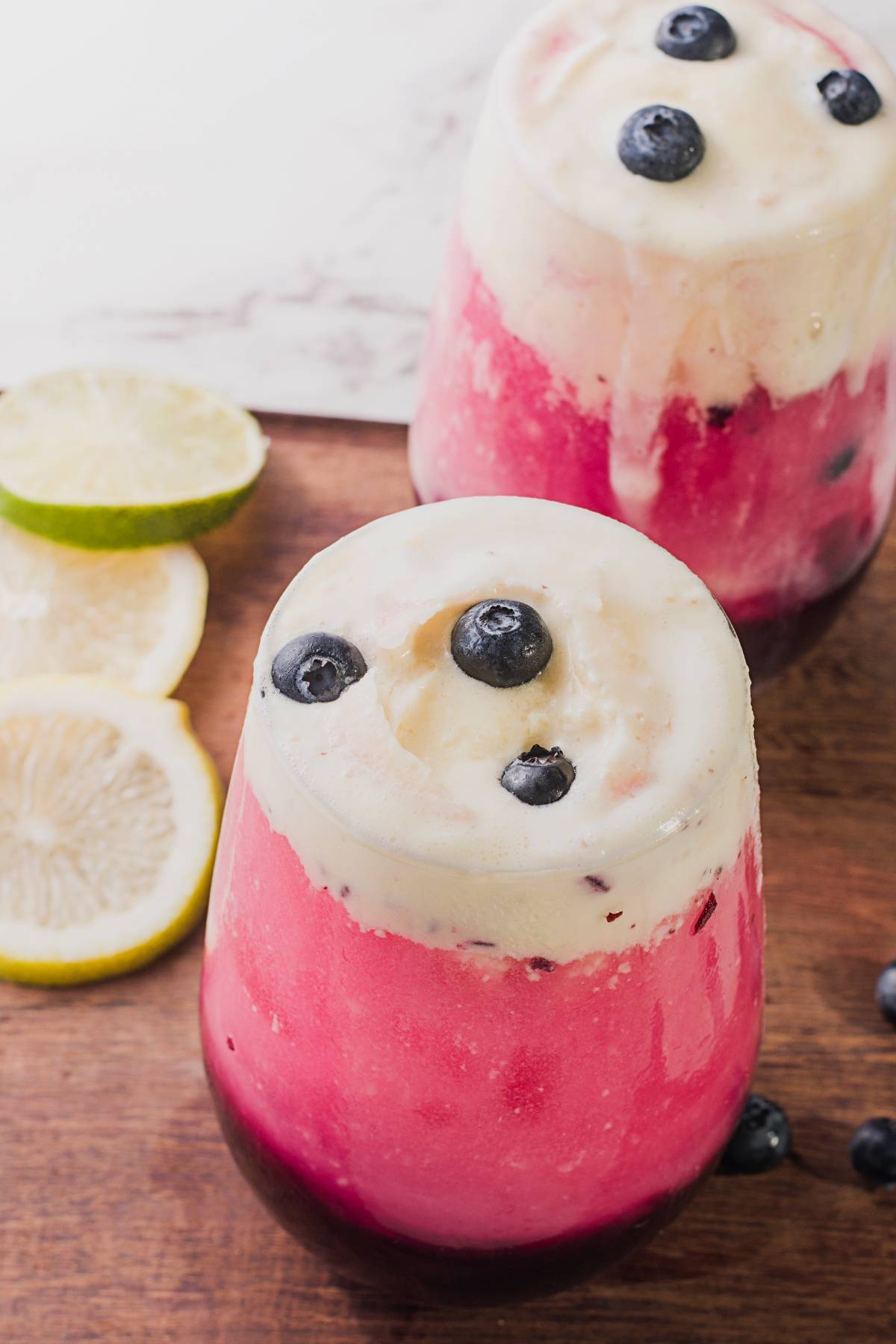 Two pink and white layered sparkling blueberry floats topped with fresh blueberries, next to lemon and lime slices on a wooden surface.
