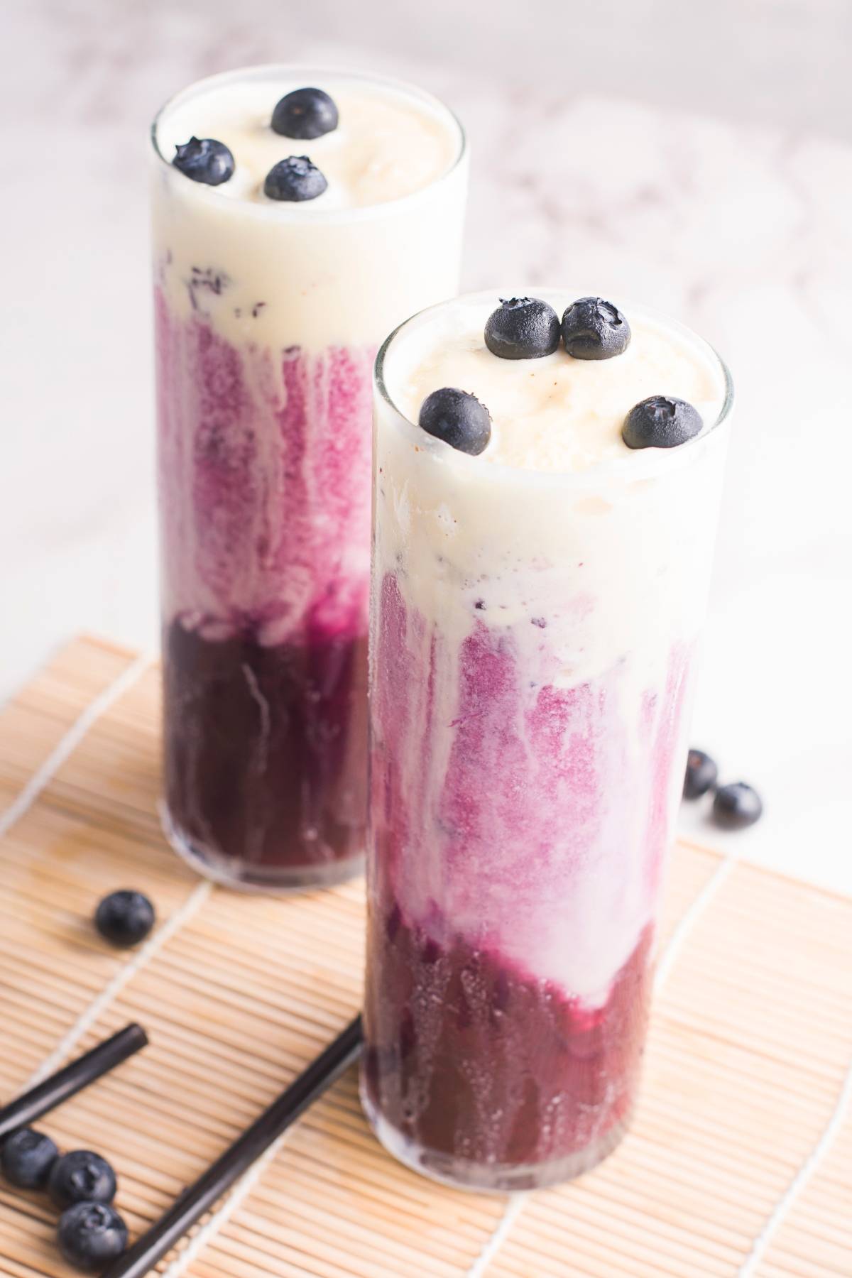 Two tall glasses of sparkling blueberry float, layered with cream and topped with blueberries, sit on a bamboo mat with straws nearby—a refreshing fresh blueberry recipe.