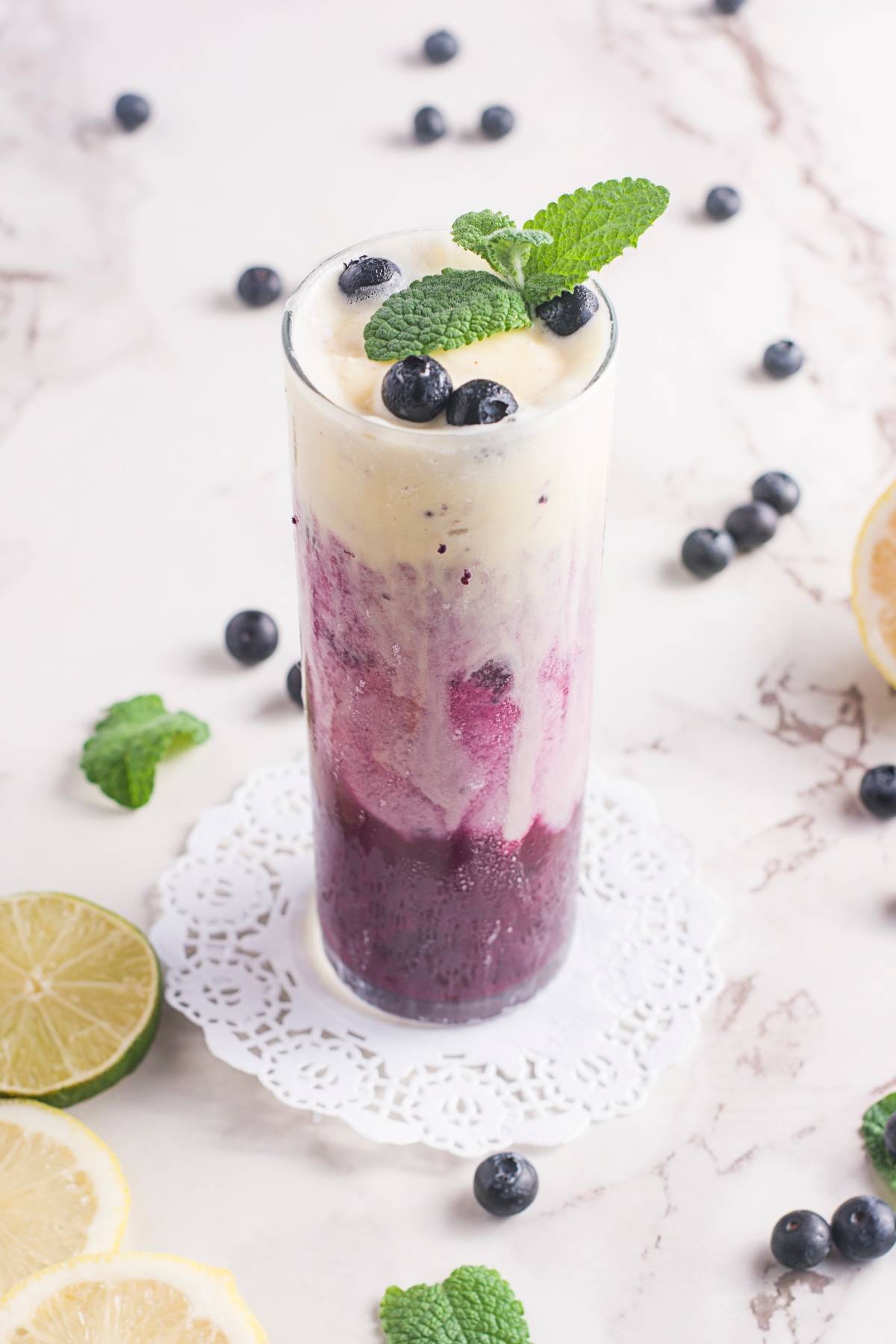 Tall glass of layered sparkling blueberry float topped with mint and blueberries, surrounded by lime slices and mint leaves—a fresh blueberry recipe perfect for summer.