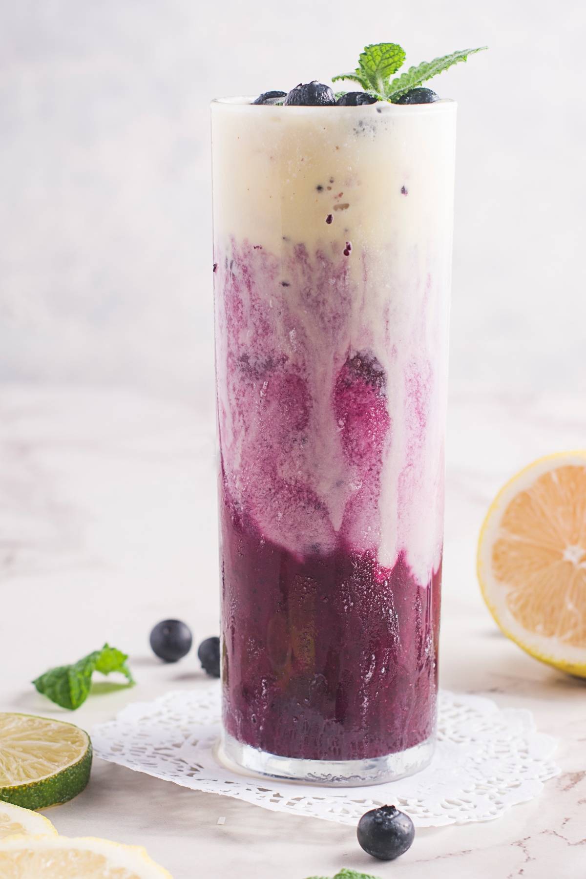 Tall glass with layered blueberry and creamy white smoothie, topped with blueberries and mint on a lace doily—a fresh blueberry recipe perfect for breakfast or a refreshing treat.