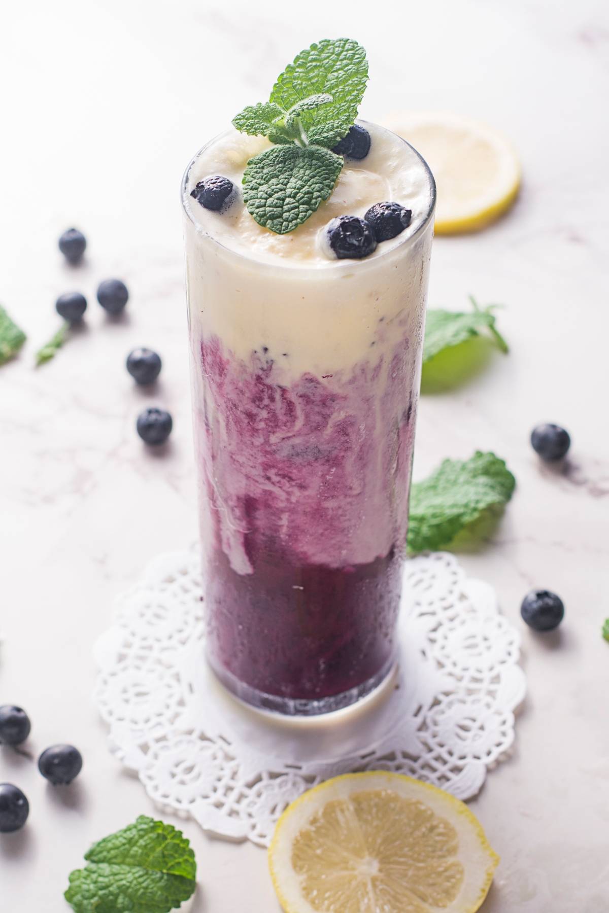 Tall glass with a layered blueberry and creamy smoothie, like a fresh blueberry recipe or sparkling blueberry float, topped with mint leaves and blueberries, on a lace doily.