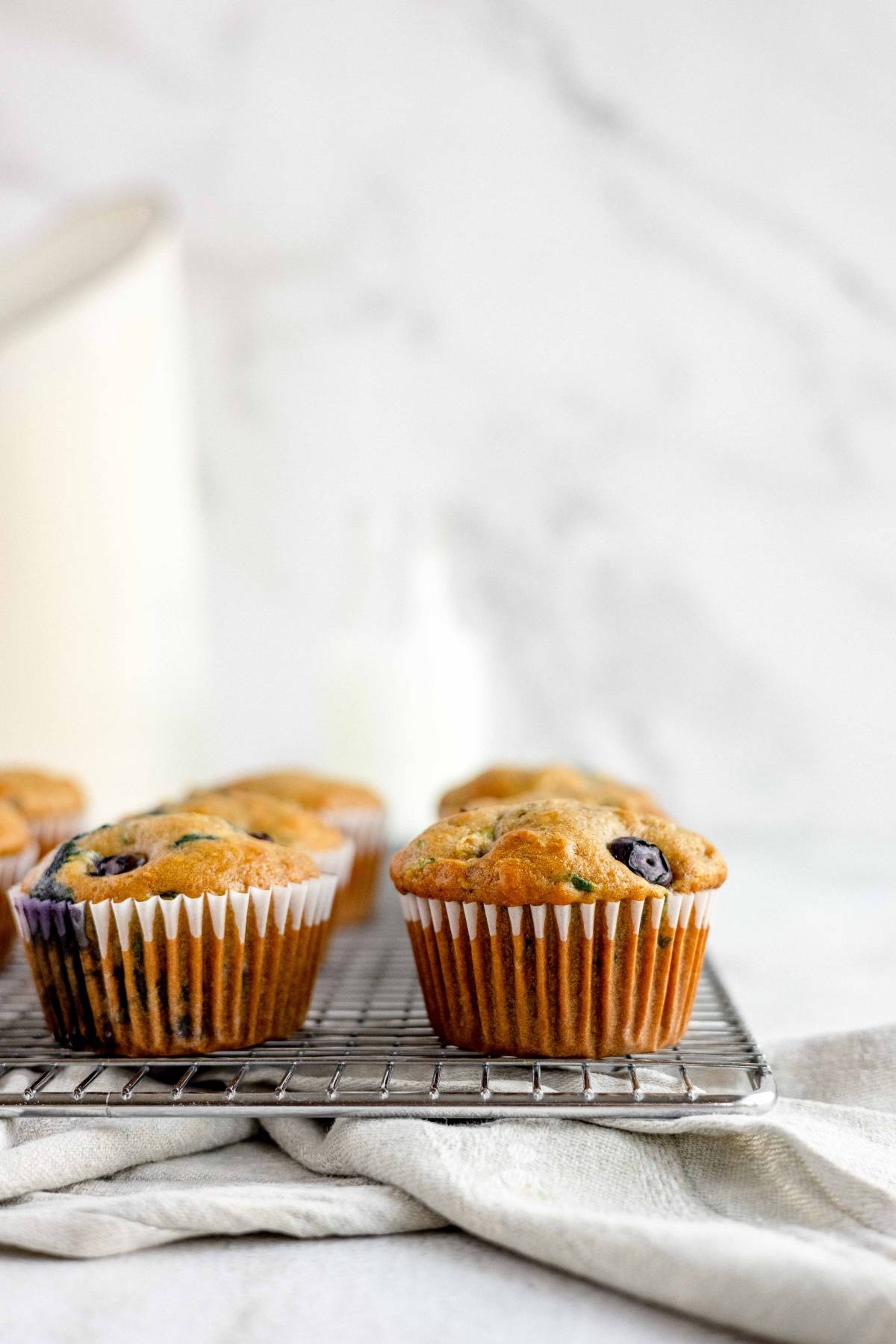 Two healthy blueberry muffins in paper liners rest on a cooling rack with a light cloth underneath, perfect for anyone seeking inspiration for their next zucchini muffin recipe.
