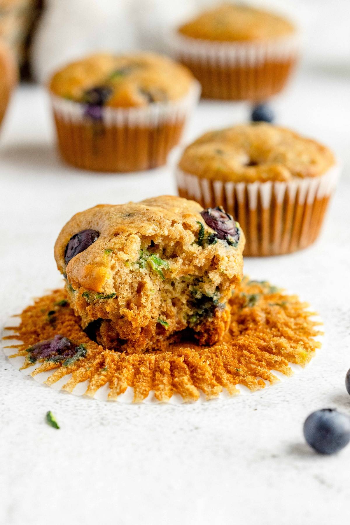 A healthy blueberry muffin with a bite taken out, sitting on an opened muffin wrapper, with whole blueberry zucchini muffins behind.