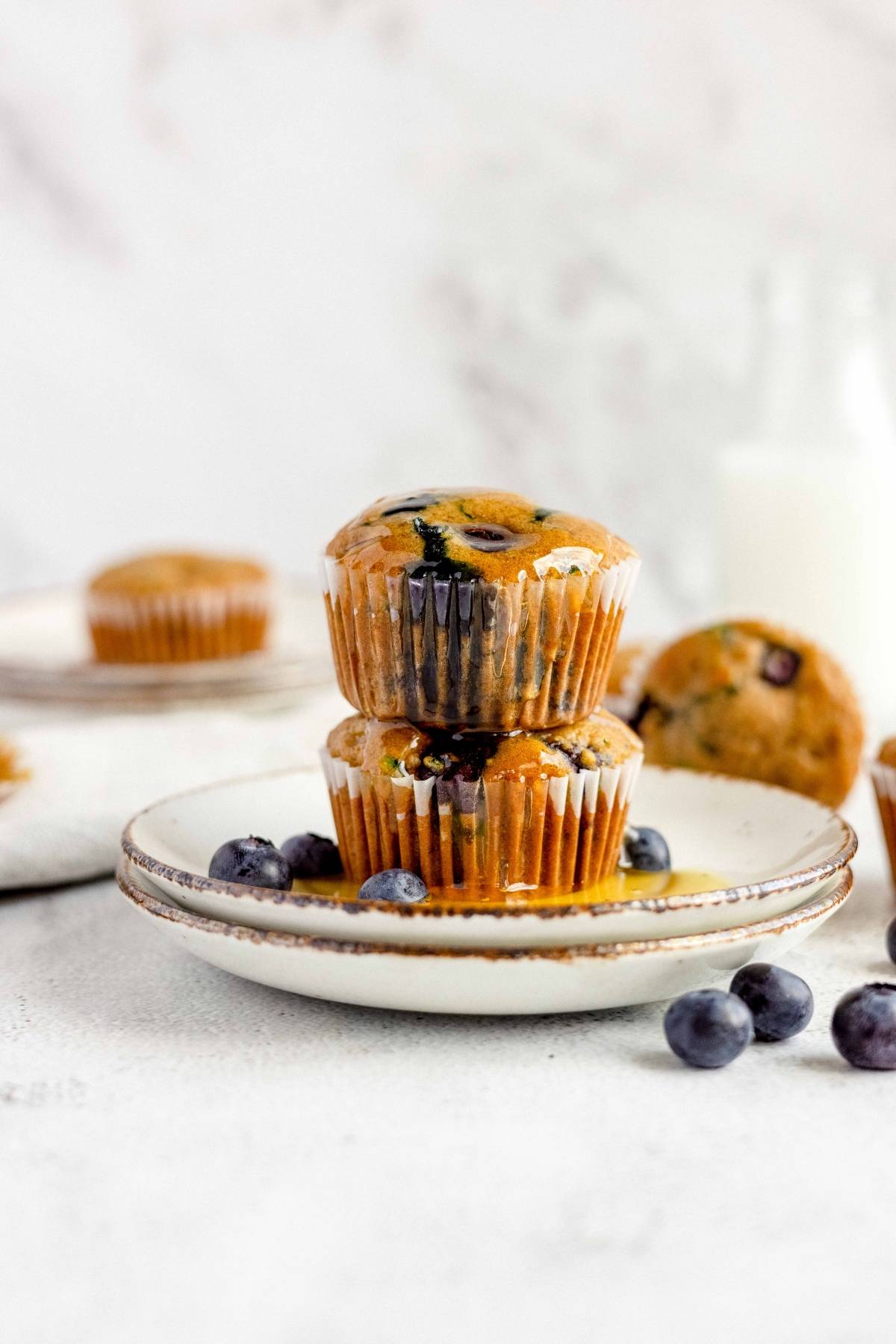 Two blueberry zucchini muffins stacked on a plate with fresh blueberries and a bottle of milk in the background.