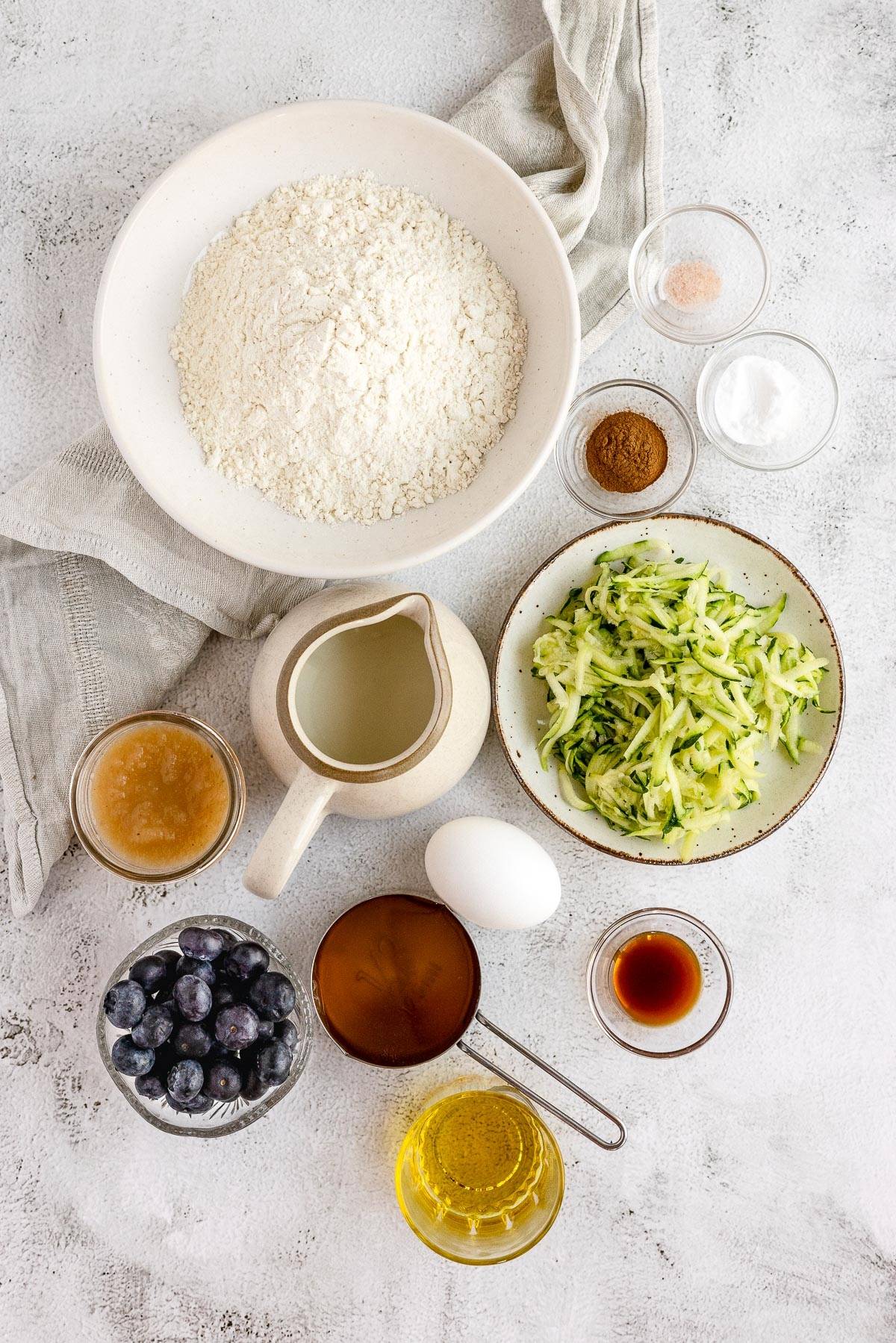 Ingredients for baking zucchini blueberry bread laid out on a white surface, perfect for a healthy blueberry muffin or zucchini muffins with applesauce, including flour, egg, and fresh blueberries.