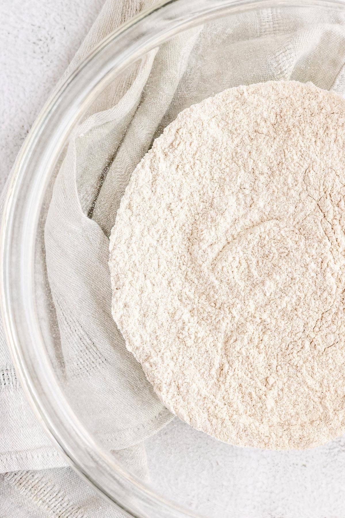 A clear bowl with flour on a light cloth, viewed from above on a light surface, ready to start a healthy blueberry muffin or zucchini muffin recipe.