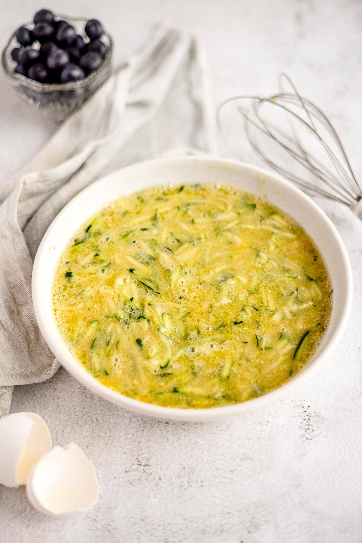 White bowl with a yellow, green-speckled egg mixture for a zucchini muffin recipe sits beside cracked eggshells, whisk, and a bowl of blueberries—perfect for healthy blueberry muffin inspiration.