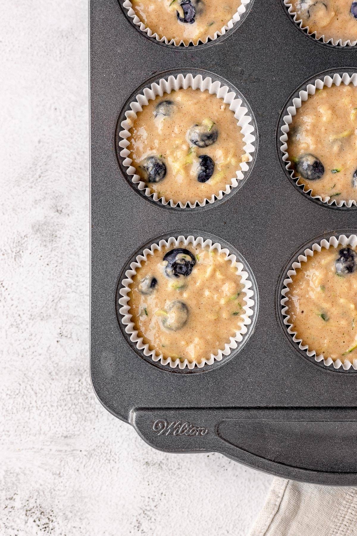 Muffin tin filled with unbaked healthy blueberry muffin batter in white paper liners on a light surface.