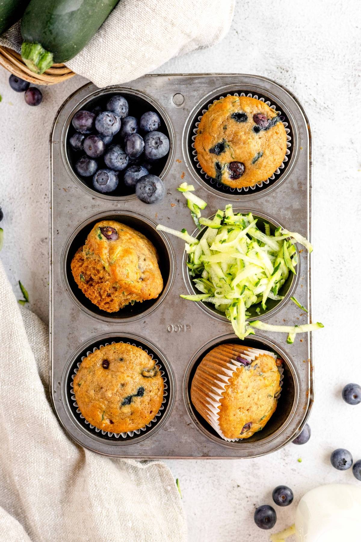 A muffin tin with healthy blueberry muffins, fresh blueberries, and shredded zucchini on a light background—perfect for trying a new zucchini muffin recipe.