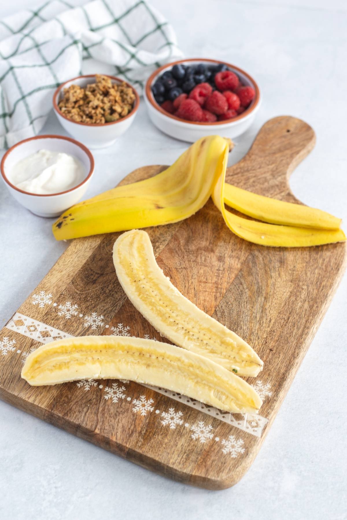 Banana halves on a wooden board create an easy no cook breakfast, with bowls of berries, granola, and yogurt in the background—perfect as an alternative to a smoothie bowl or as a Breakfast Banana Split.
