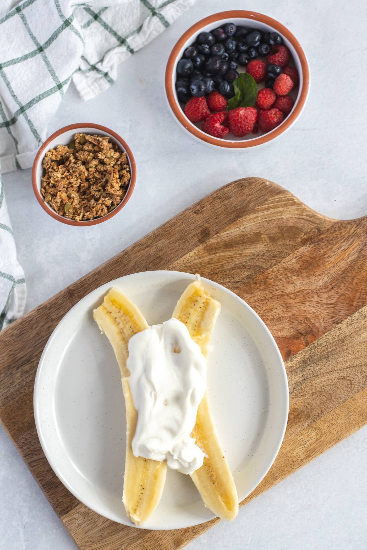 A banana split with yogurt on a plate, next to bowls of granola and banana and fresh berries—an easy no-cook breakfast that's a tasty alternative to a smoothie bowl.