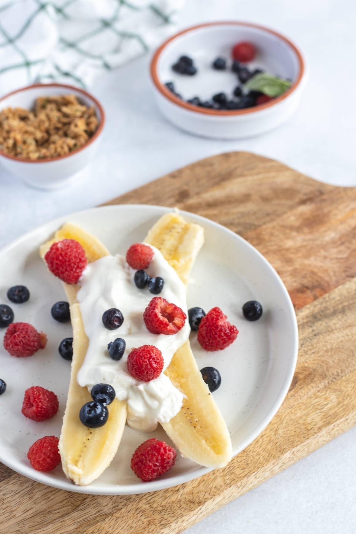 A Breakfast Banana Split with banana and fresh berries, topped with yogurt, raspberries, and blueberries on a white plate—one of the best healthy breakfast ideas for kids. Granola bowls sit nearby for an added crunch.