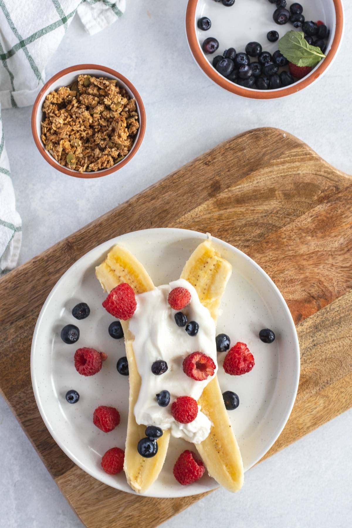 A Breakfast Banana Split topped with whipped cream, raspberries, and blueberries sits on a plate beside bowls of granola and blueberries—one of the best healthy breakfast ideas for kids who love banana and fresh berries.