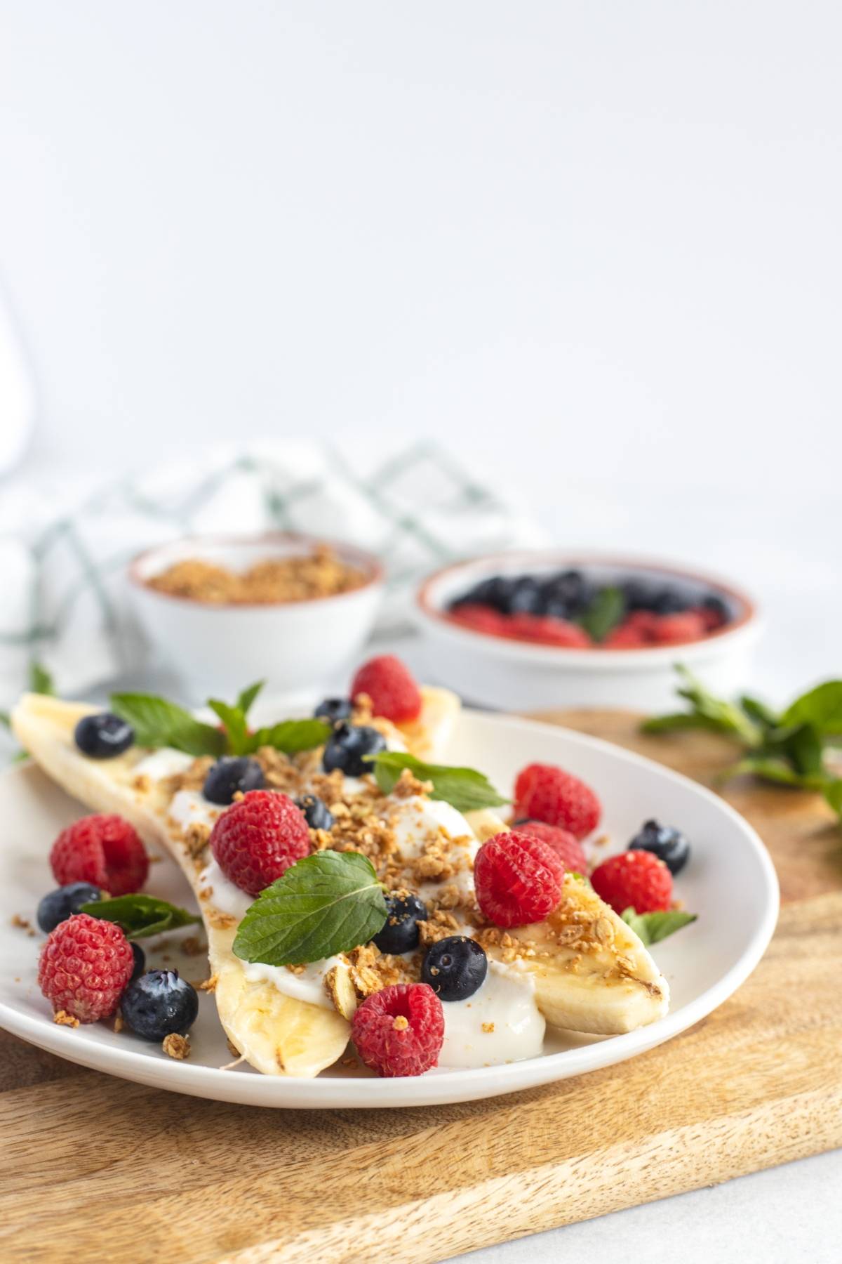 A Breakfast Banana Split topped with yogurt, granola, raspberries, blueberries, and mint leaves on a white plate—an easy no cook breakfast and one of the best healthy breakfast ideas for kids.