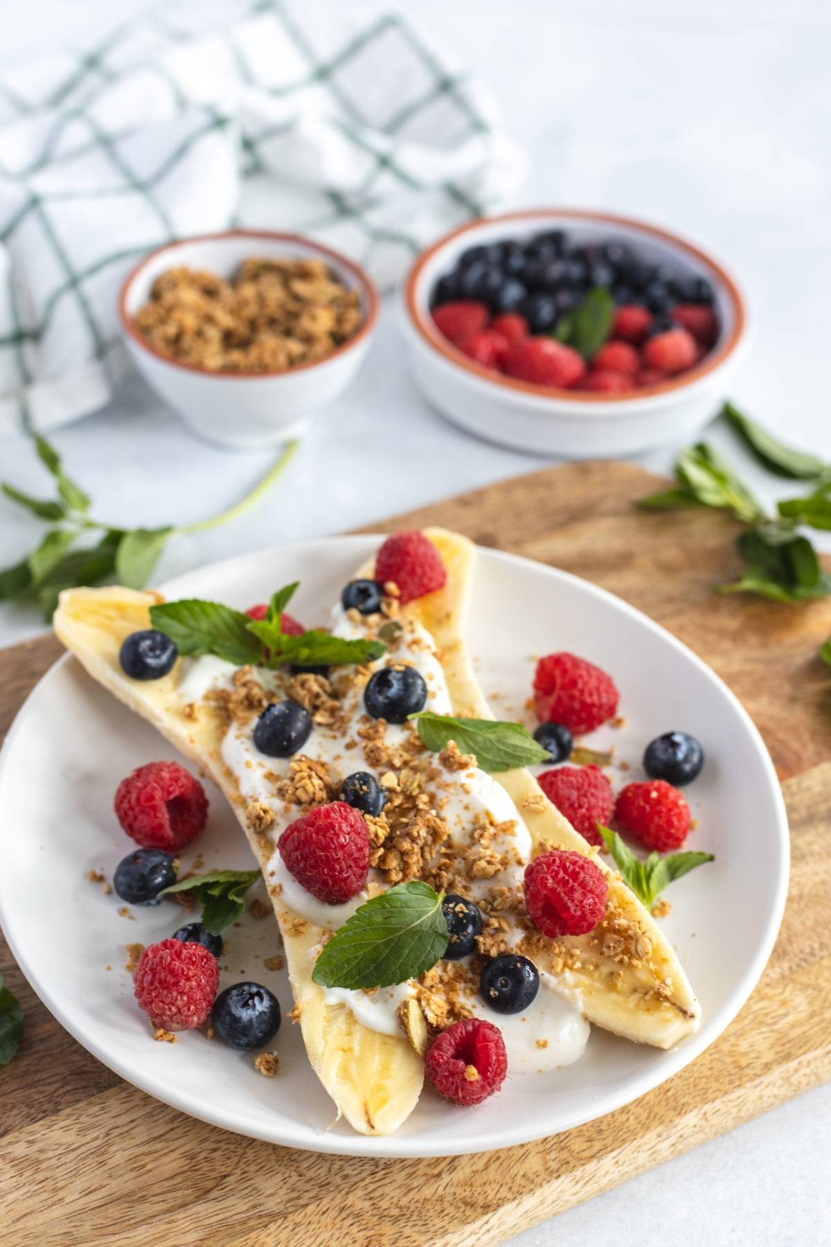 A banana split with yogurt, granola, banana and fresh berries, plus mint on a white plate—one of the best healthy breakfast ideas for kids and an easy no cook breakfast everyone will love.