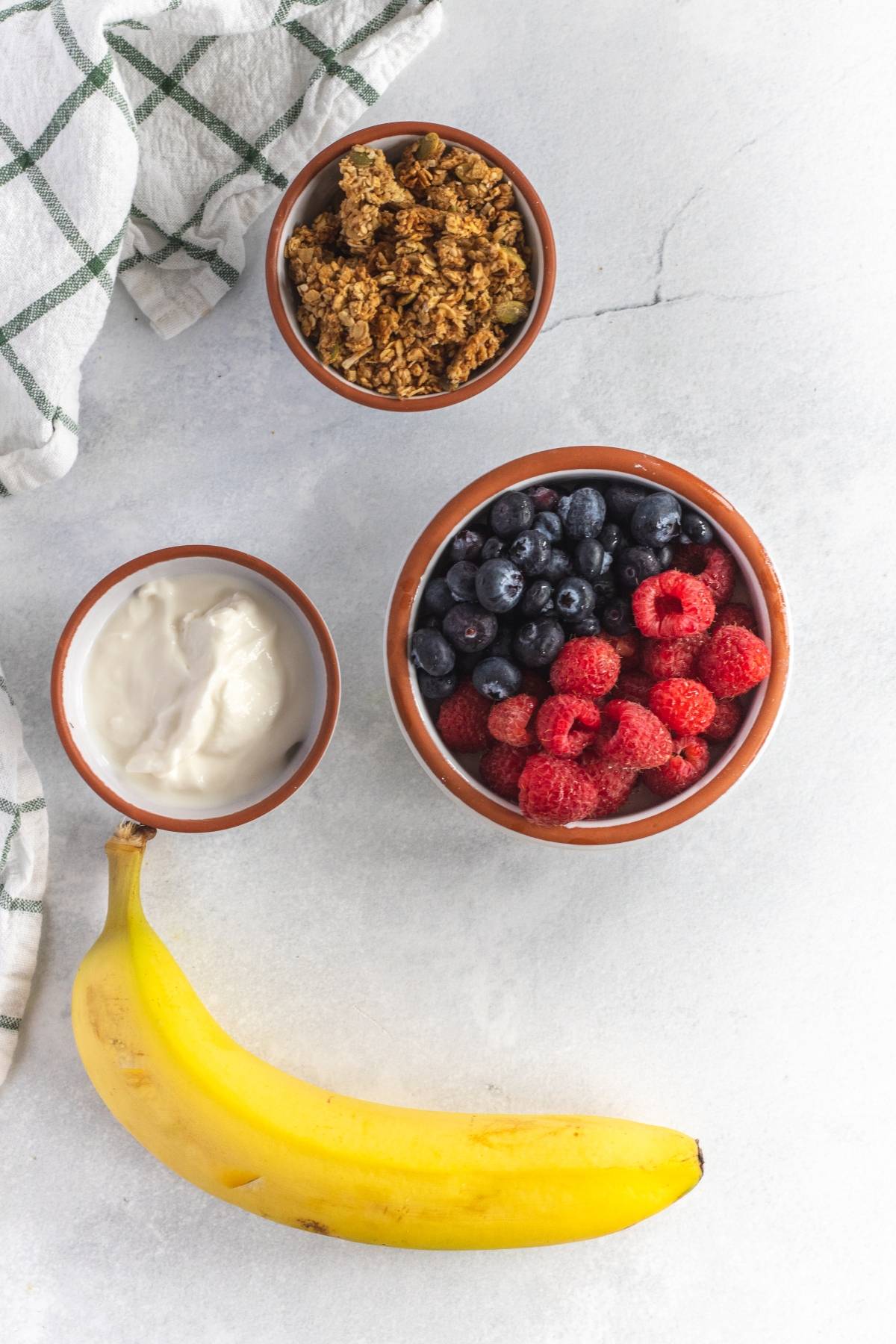 A banana, yogurt, granola, and a bowl of raspberries and blueberries on a light surface—an easy no cook breakfast and a great alternative to a smoothie bowl for healthy breakfast ideas for kids.