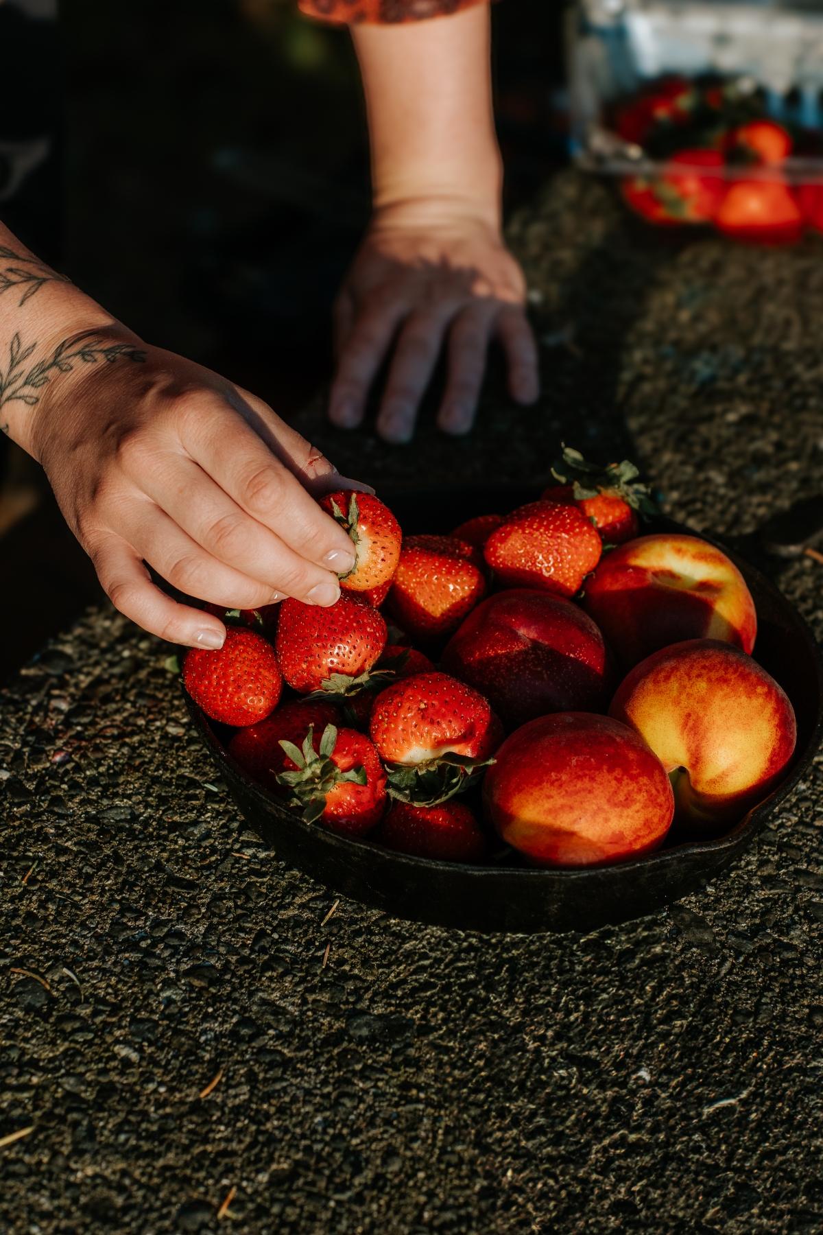 A hand picks a strawberry from a bowl filled with strawberries and peaches on a textured surface, perfect ingredients for a fresh campfire cobbler.