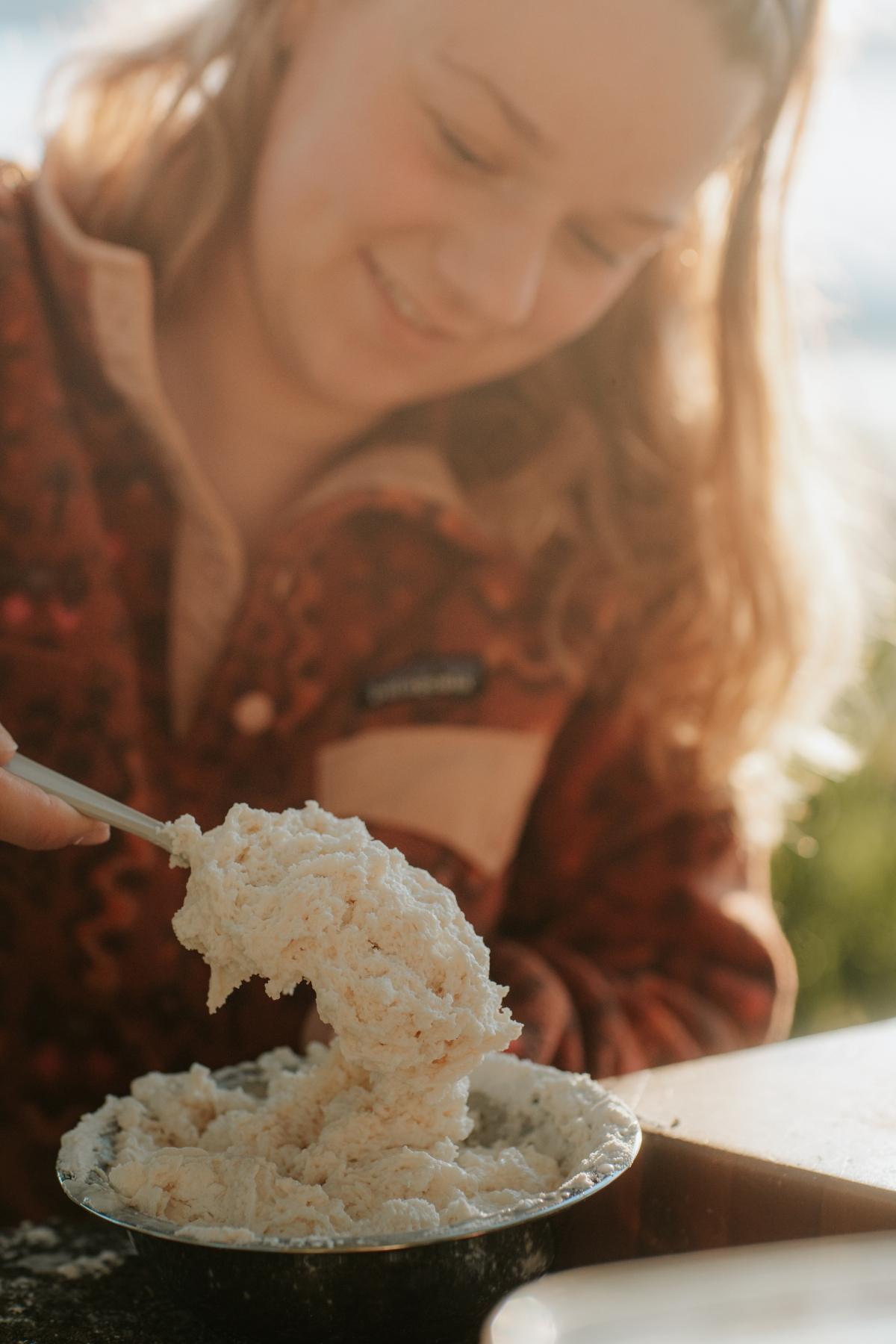 A person smiles while scooping a bowl of creamy, textured spread outdoors in sunlight, the aroma of campfire cobbler lingering in the fresh air.
