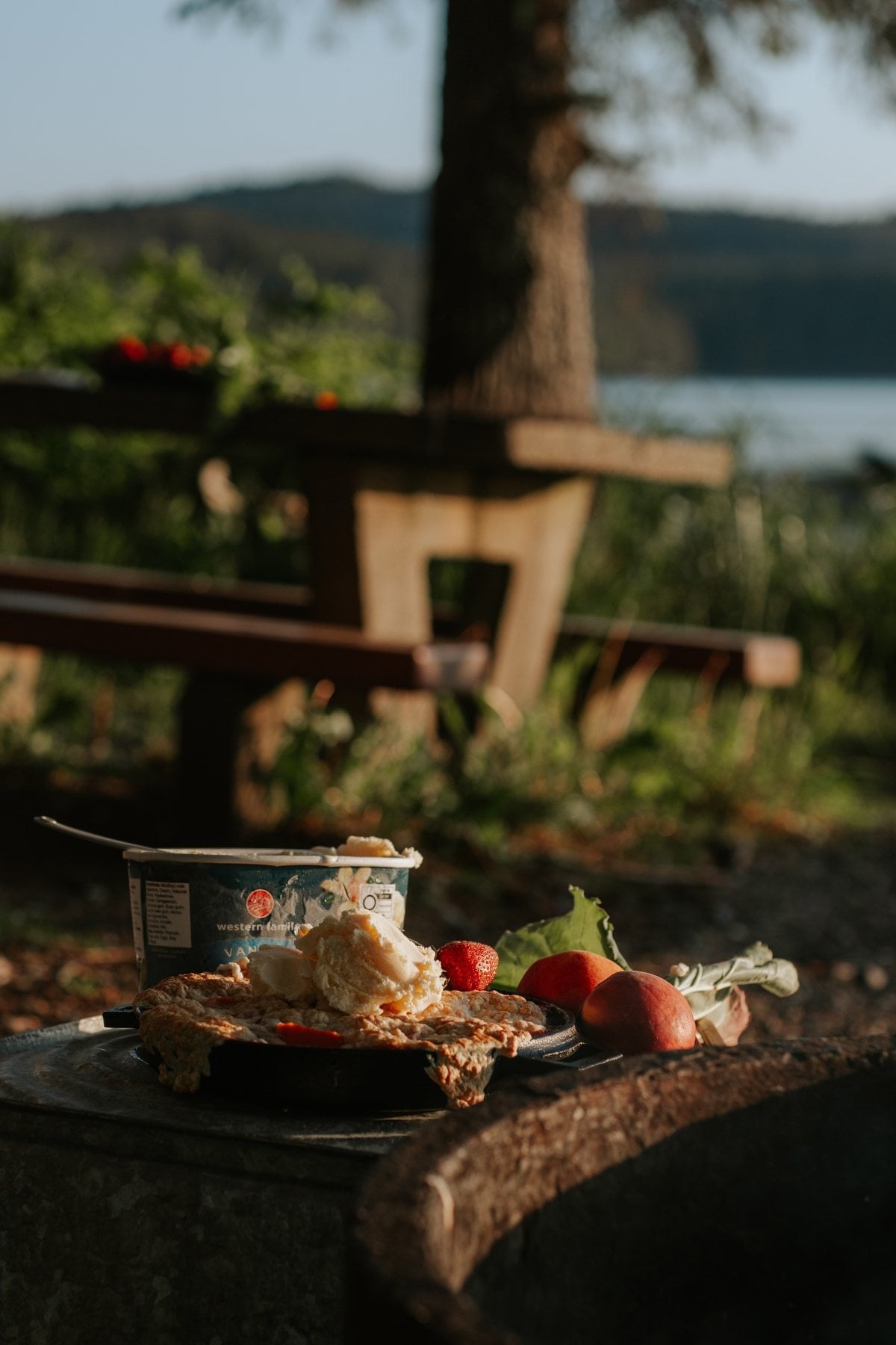 A picnic table by a lake with pie, strawberries, cream, and campfire cobbler in the soft sunlight.