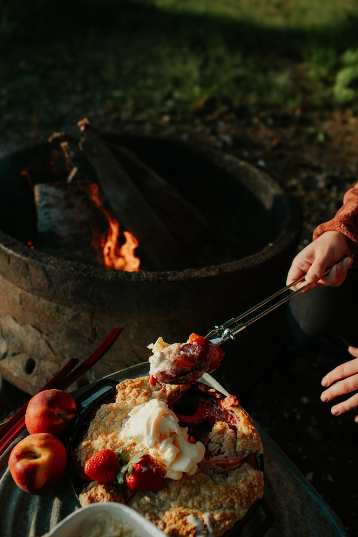 A person serves campfire cobbler with ice cream near a campfire, with peaches and strawberries beside the dish.