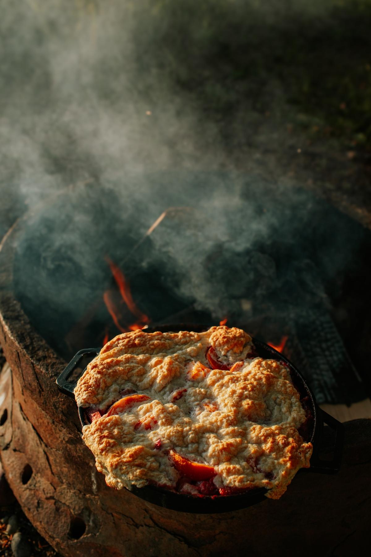 A campfire cobbler dessert baking in a cast iron pan over a smoky outdoor fire pit.