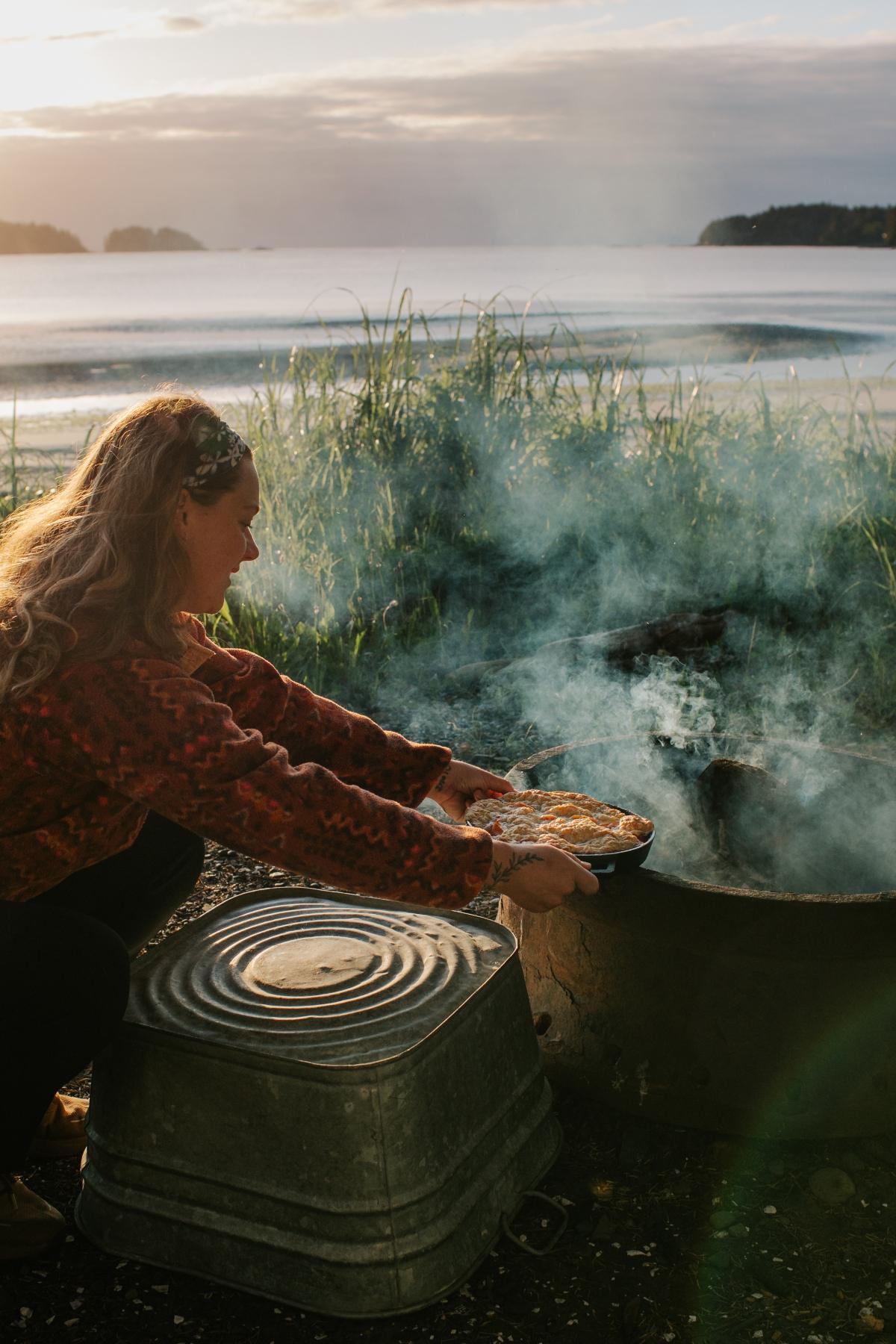 A woman cooks campfire cobbler over a smoky fire by a lake at sunset, holding a pan filled with delicious food.