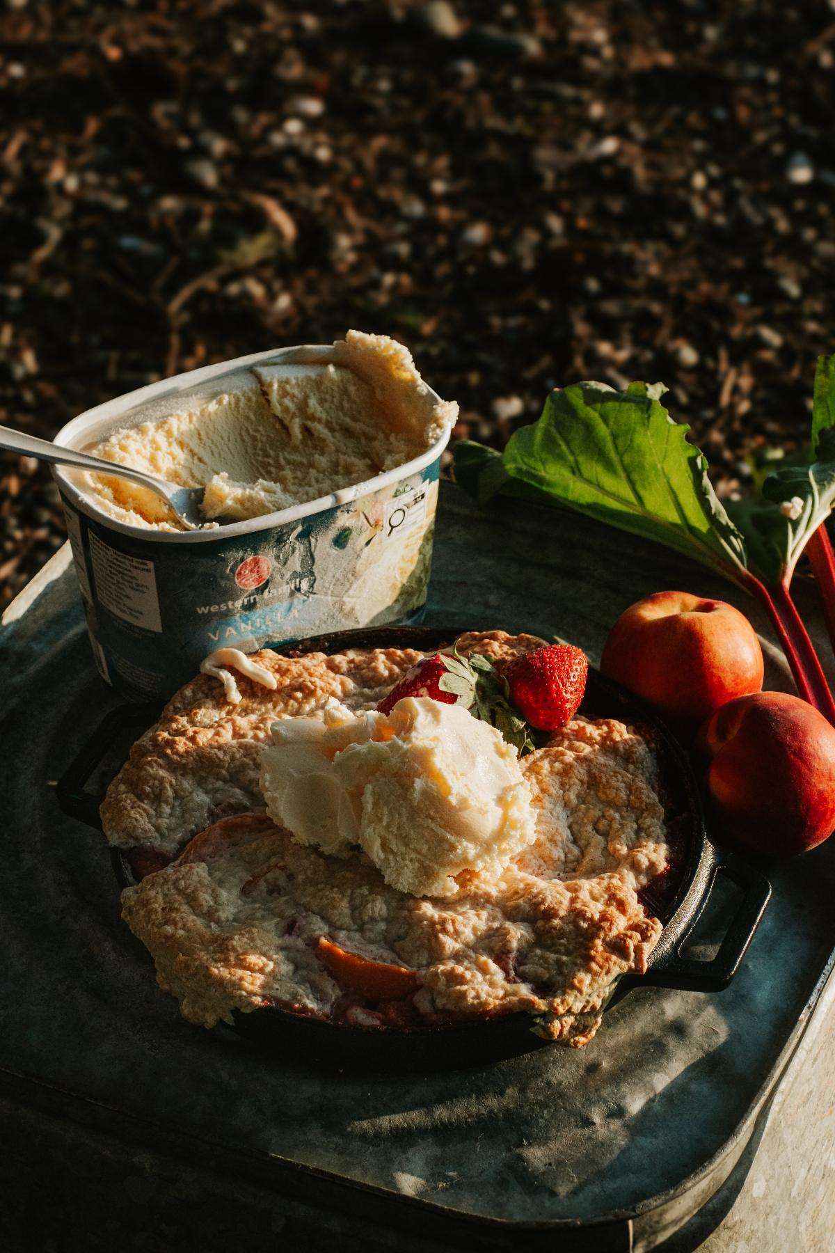 A campfire cobbler topped with vanilla ice cream sits beside fresh peaches and a tub of ice cream outdoors.