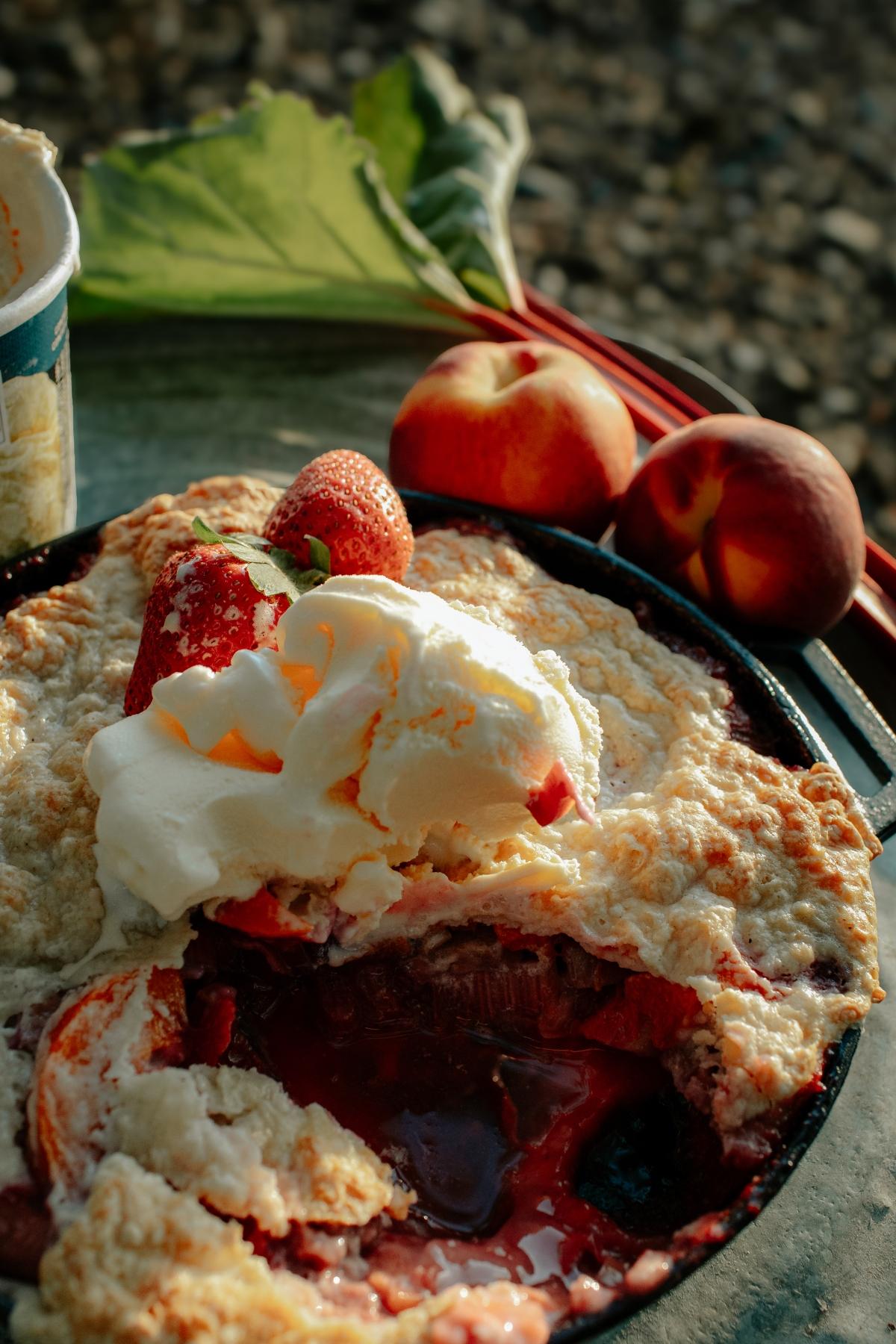 A campfire cobbler fruit pie topped with vanilla ice cream, surrounded by peaches and strawberries on a table.