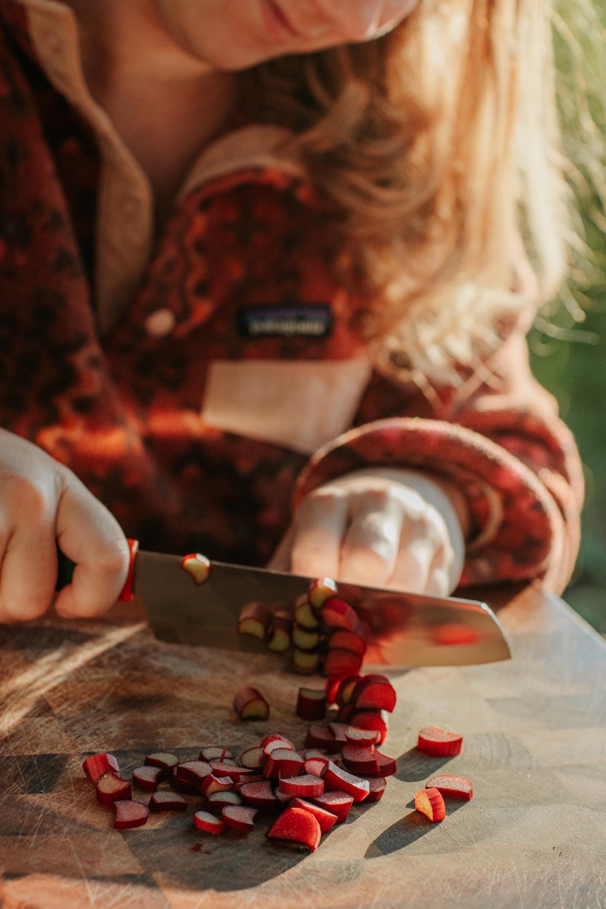 Person chopping red vegetable pieces on a wooden cutting board in sunlight, preparing ingredients for a hearty campfire cobbler.