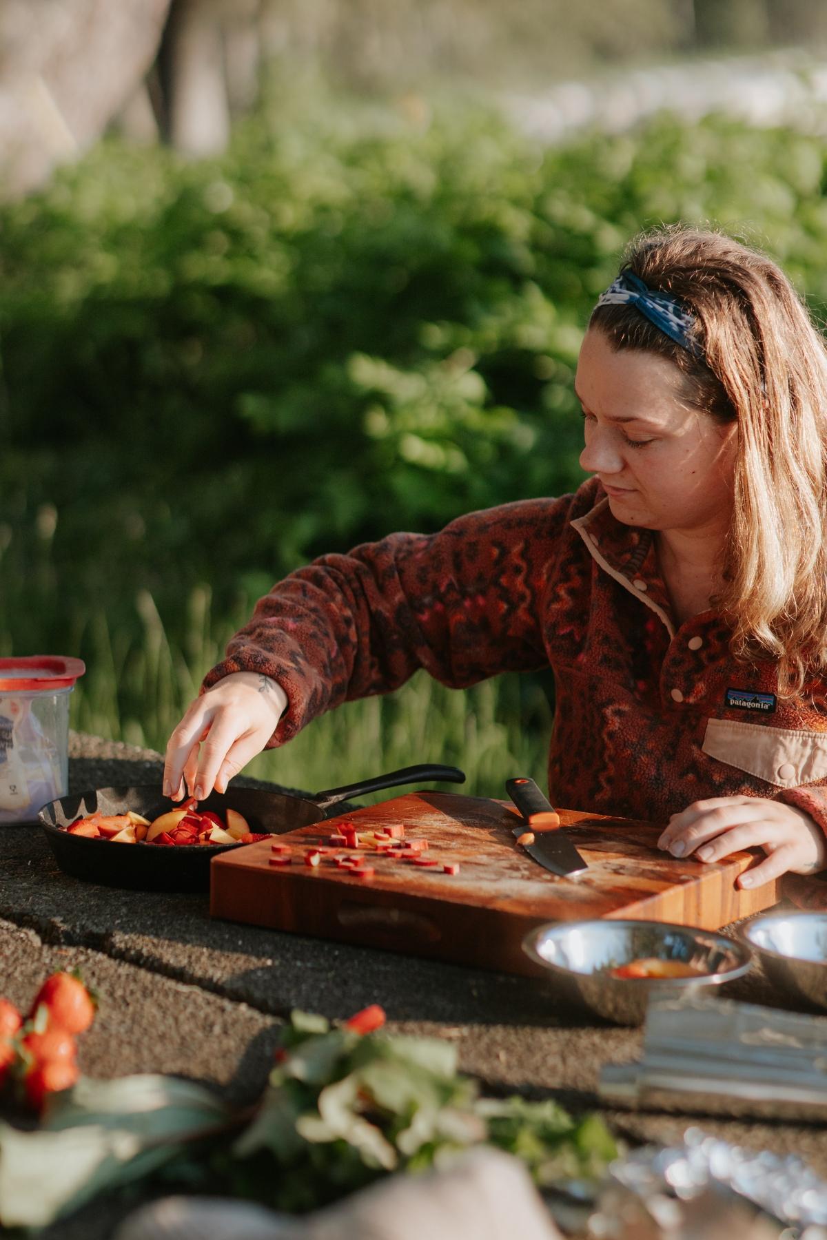 A woman slices strawberries on a wooden cutting board at an outdoor picnic table, preparing fresh fruit for a delicious campfire cobbler.