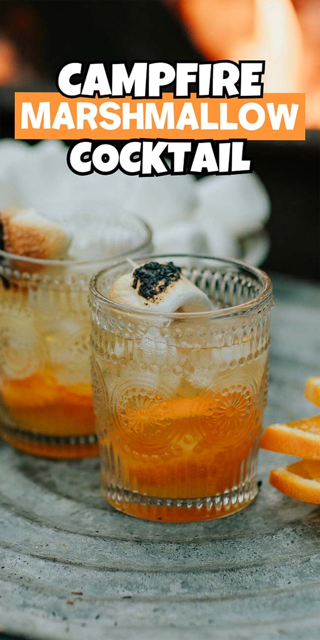 Two glasses of campfire marshmallow cocktail, topped with toasted marshmallows, sit on a stone surface with orange slices nearby.