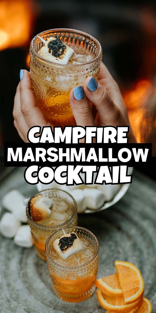 Two glasses of campfire marshmallow cocktail with toasted marshmallows, ice, and orange slices, held over a rustic table.