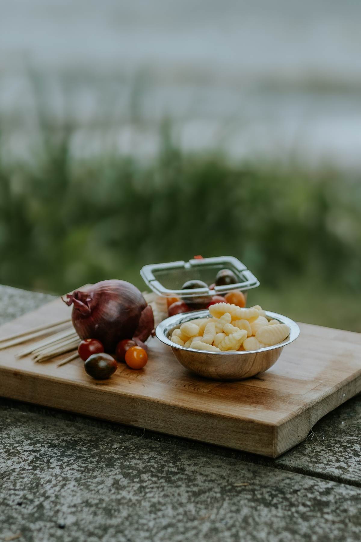 A bowl of gnocchi, cherry tomatoes, and a red onion sits on a wooden cutting board outdoors, ready to be transformed into Campfire Gnocchi Kebabs.