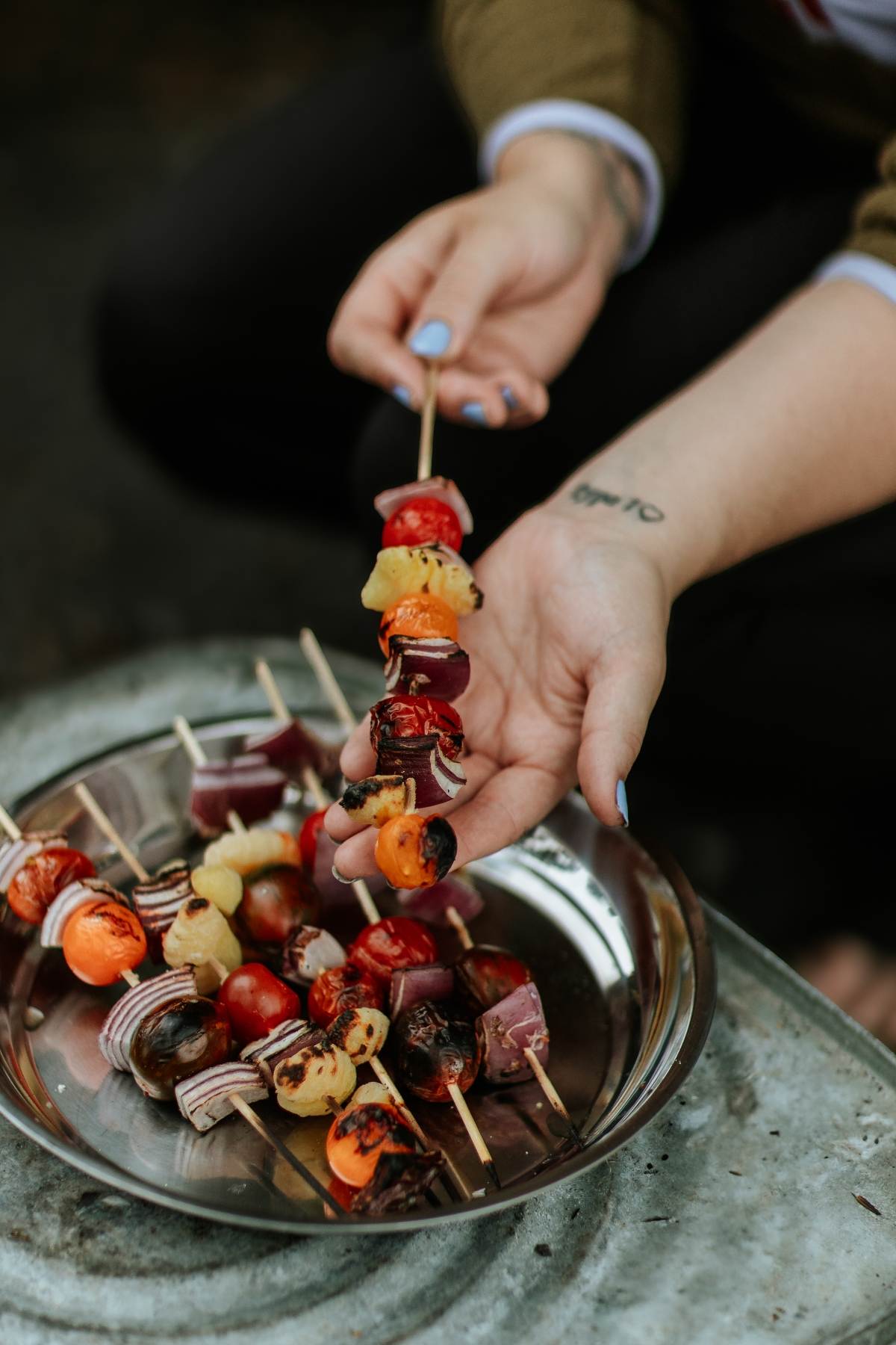 A person holds a skewer of grilled vegetables over a plate piled with more veggie skewers, creating the perfect campfire gnocchi kebabs experience.