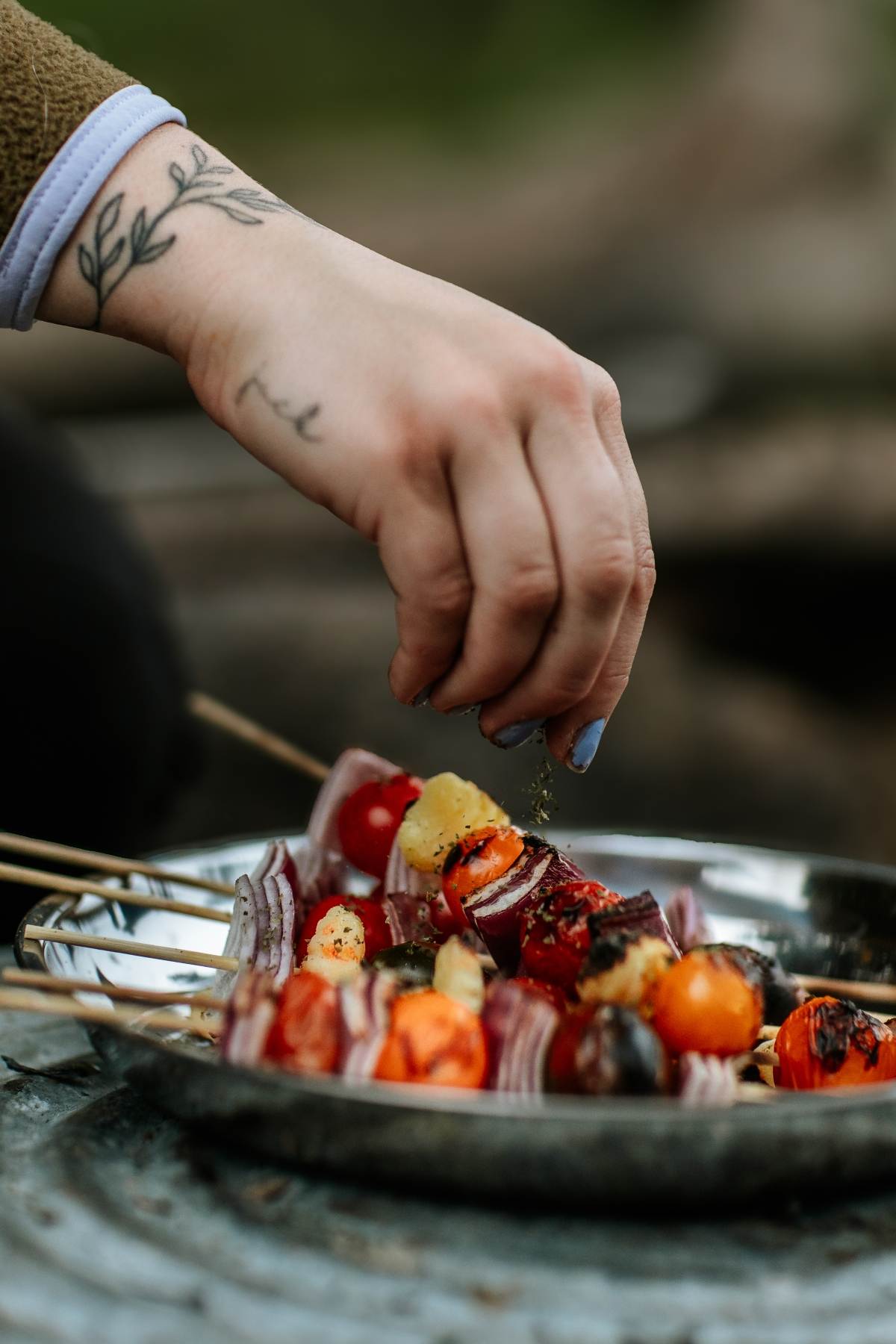A hand sprinkles seasoning on skewers of vegetables, including tomatoes and onions, for delicious Campfire Gnocchi Kebabs on a metal plate.