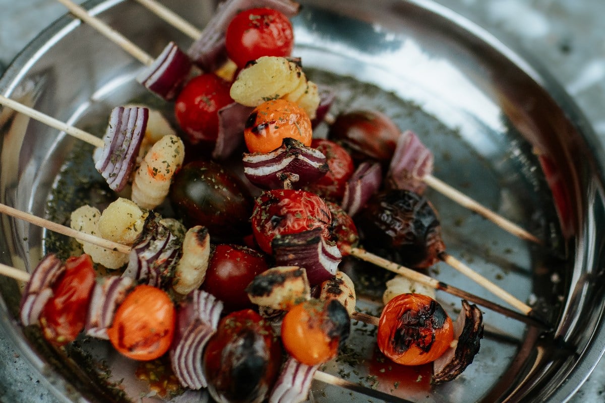 Campfire Gnocchi Kebabs featuring grilled vegetable skewers with tomatoes, onions, and potatoes on a metal plate.
