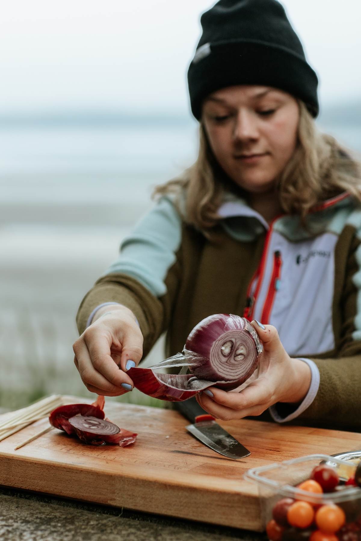 A woman in a beanie slices a red onion on a wooden board outdoors near the water, prepping ingredients for Campfire Gnocchi Kebabs.