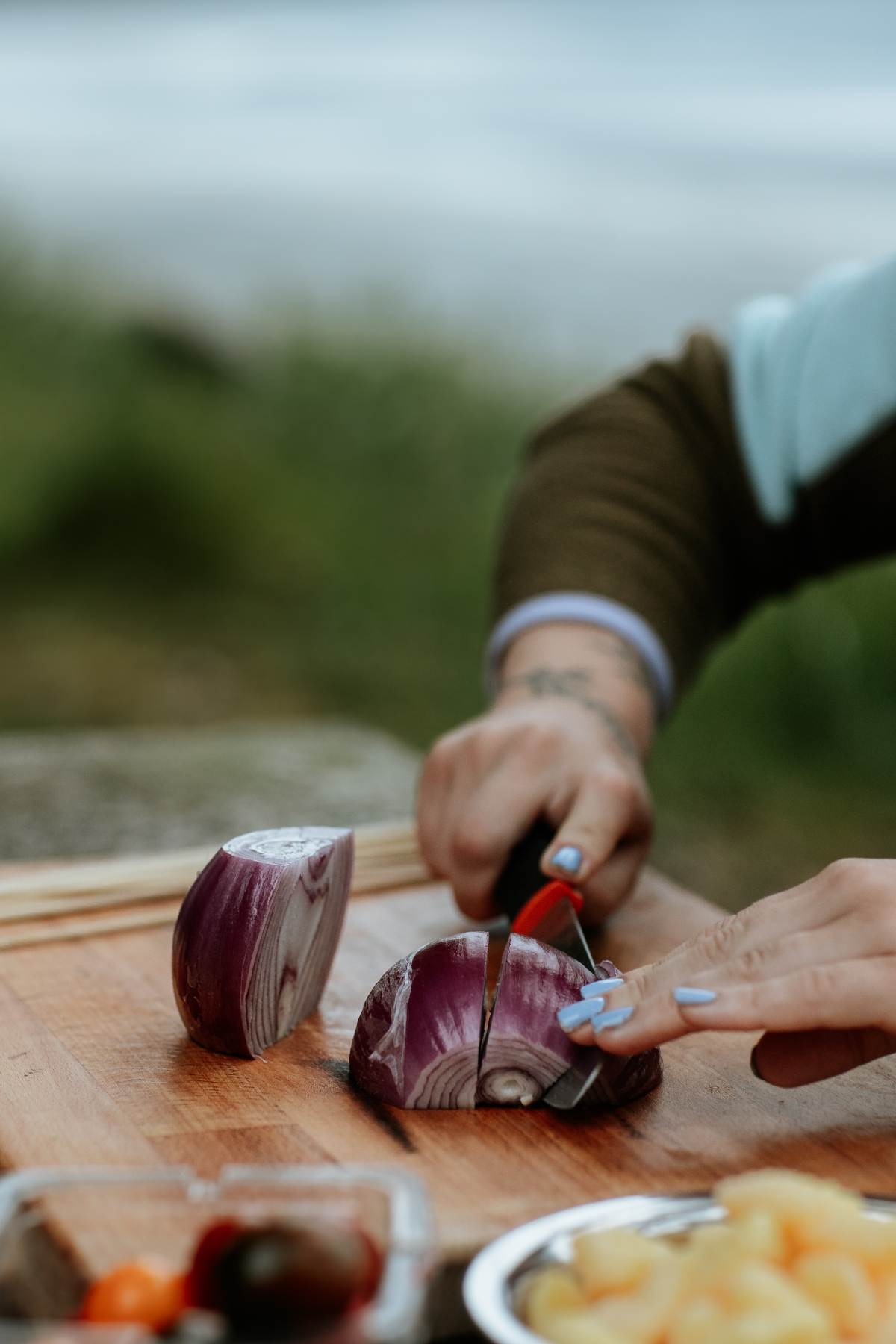 A person slices a red onion on a wooden board outdoors, preparing fresh ingredients for Campfire Gnocchi Kebabs, with other food items nearby.