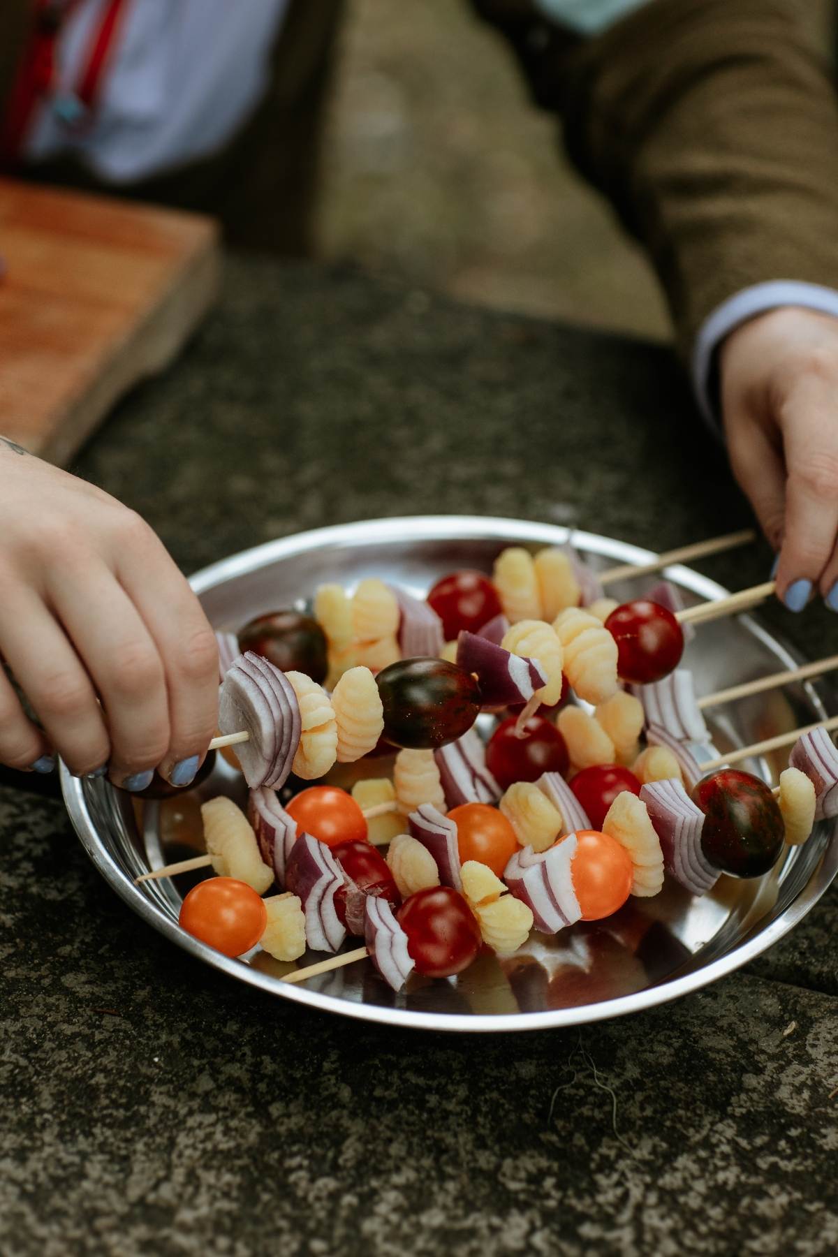 A person holding Campfire Gnocchi Kebabs with cherry tomatoes, onions, and pasta skewered over a metal bowl.