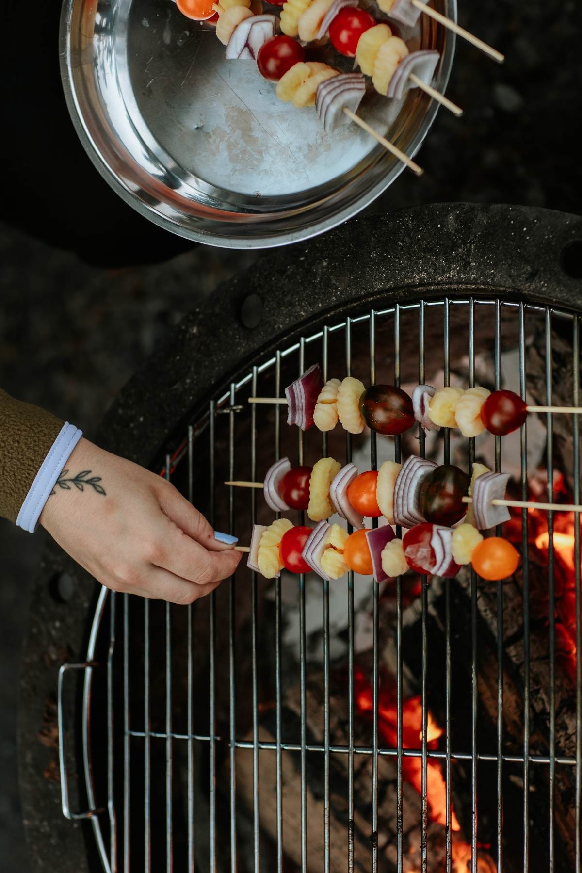 Hand placing Campfire Gnocchi Kebabs on a grill over an open flame, with more skewers on a metal plate above.