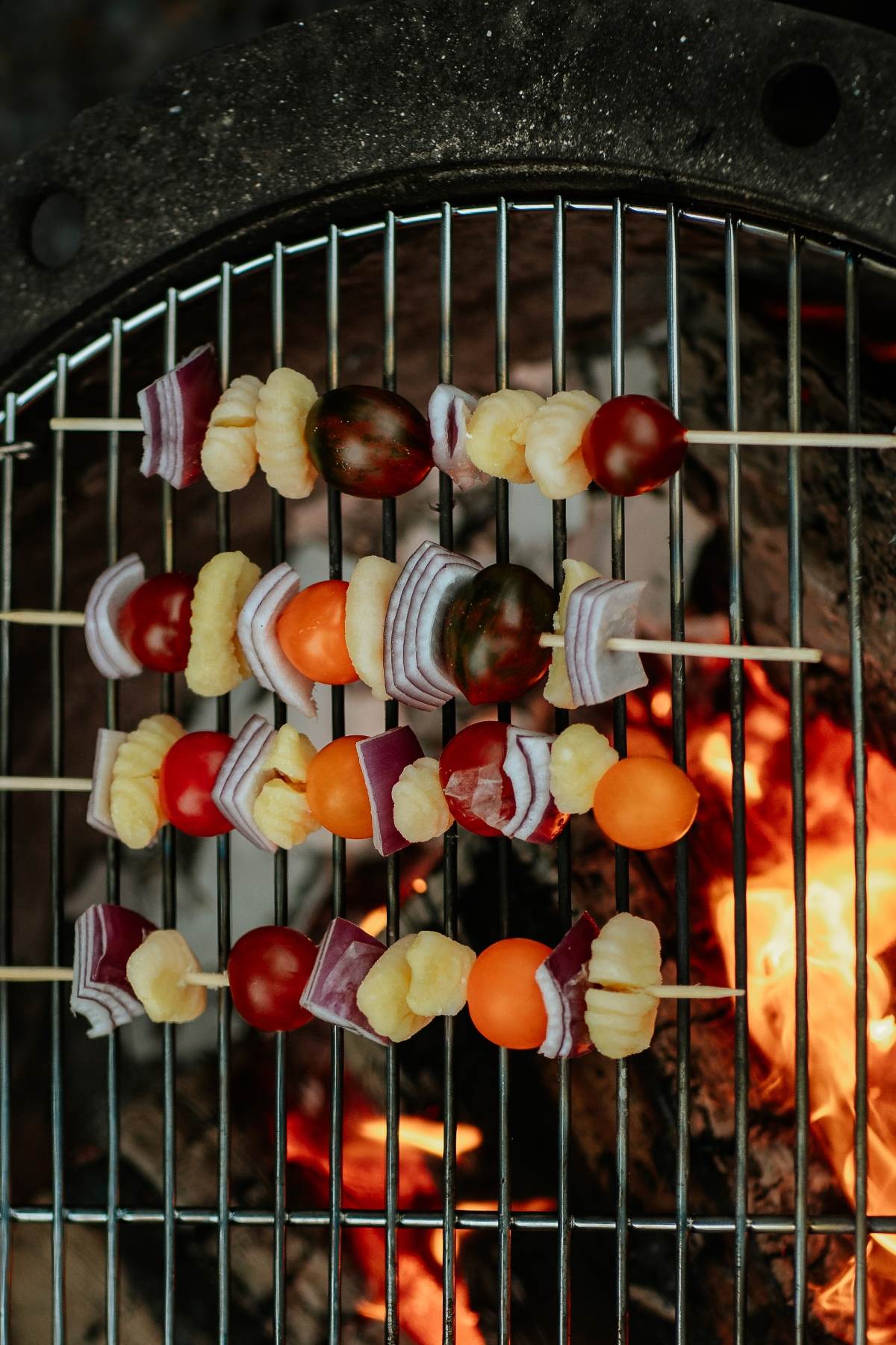 Campfire Gnocchi Kebabs sizzle alongside tomatoes, onions, and cauliflower as they grill over an open flame on a barbecue.