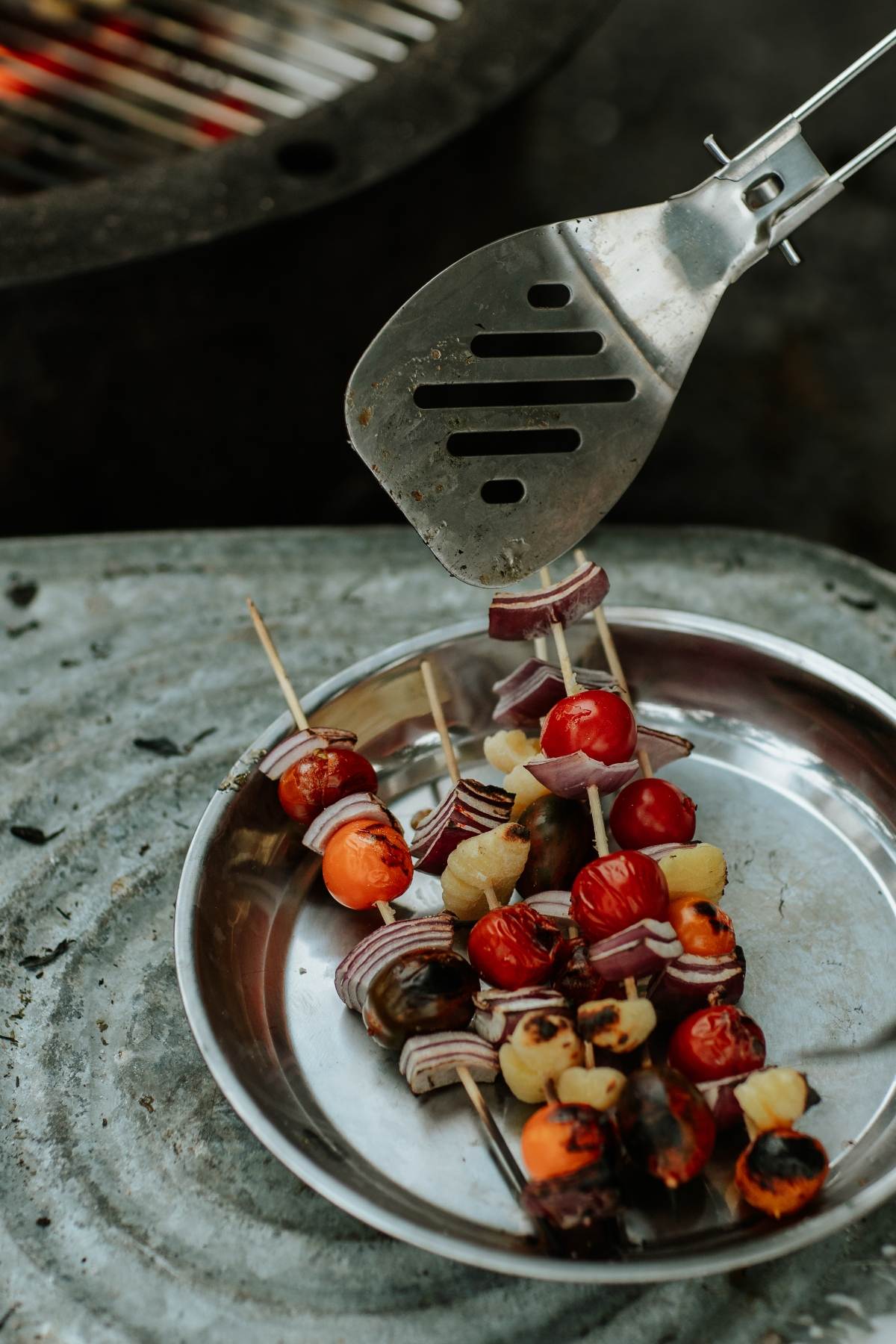 Grilled vegetable skewers with tomatoes, onions, and peppers on a metal plate, reminiscent of tasty Campfire Gnocchi Kebabs, picked up with a spatula.