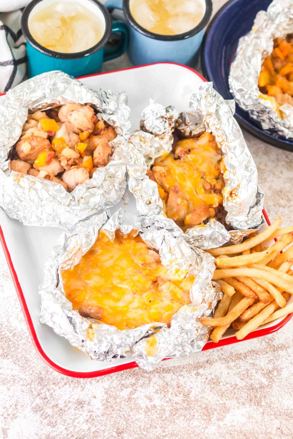 Foil packets with cheesy chicken, served with French fries, and mugs of coffee in the background.