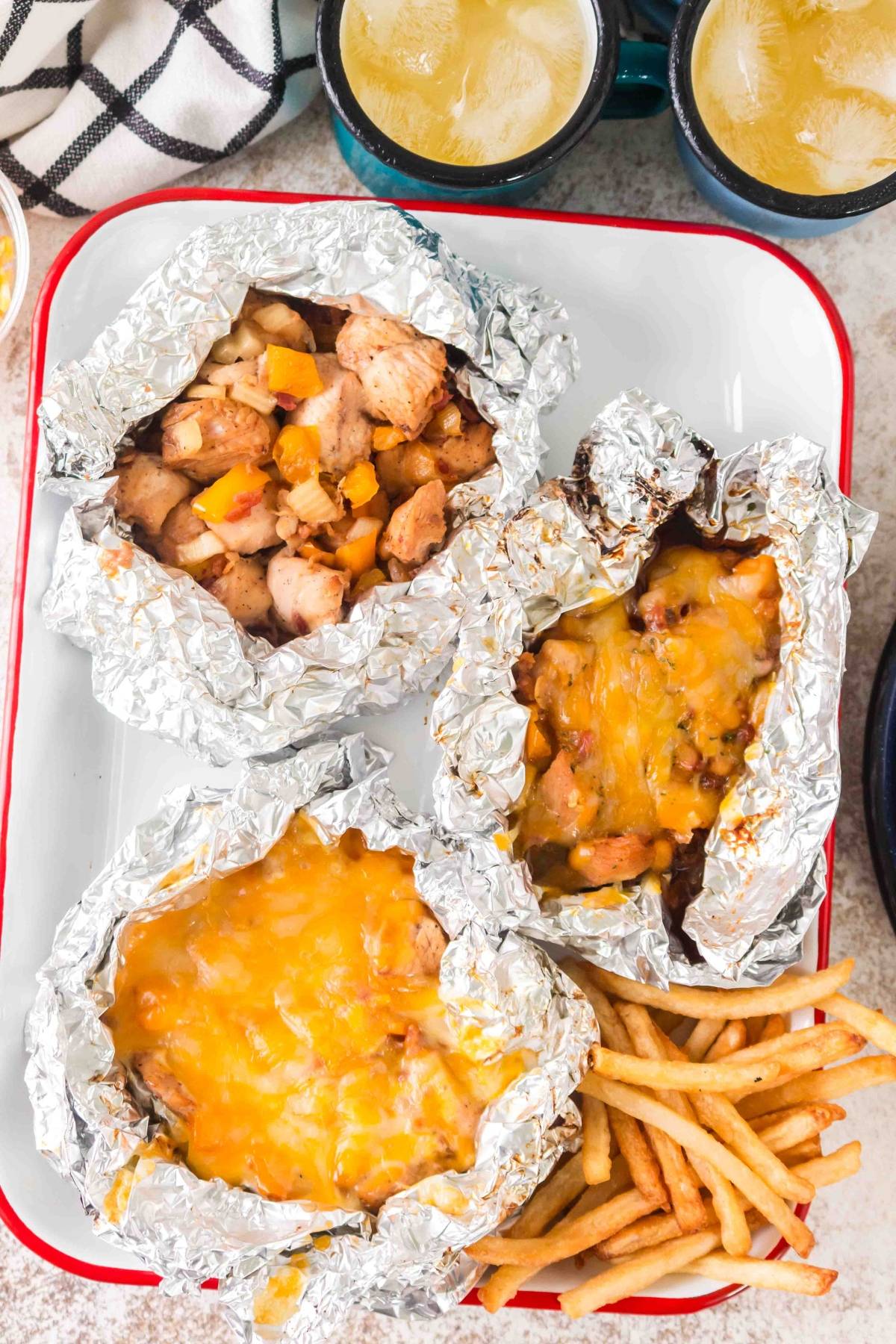 Three foil packets with cheesy meat and veggies, served with French fries and drinks on a tray.