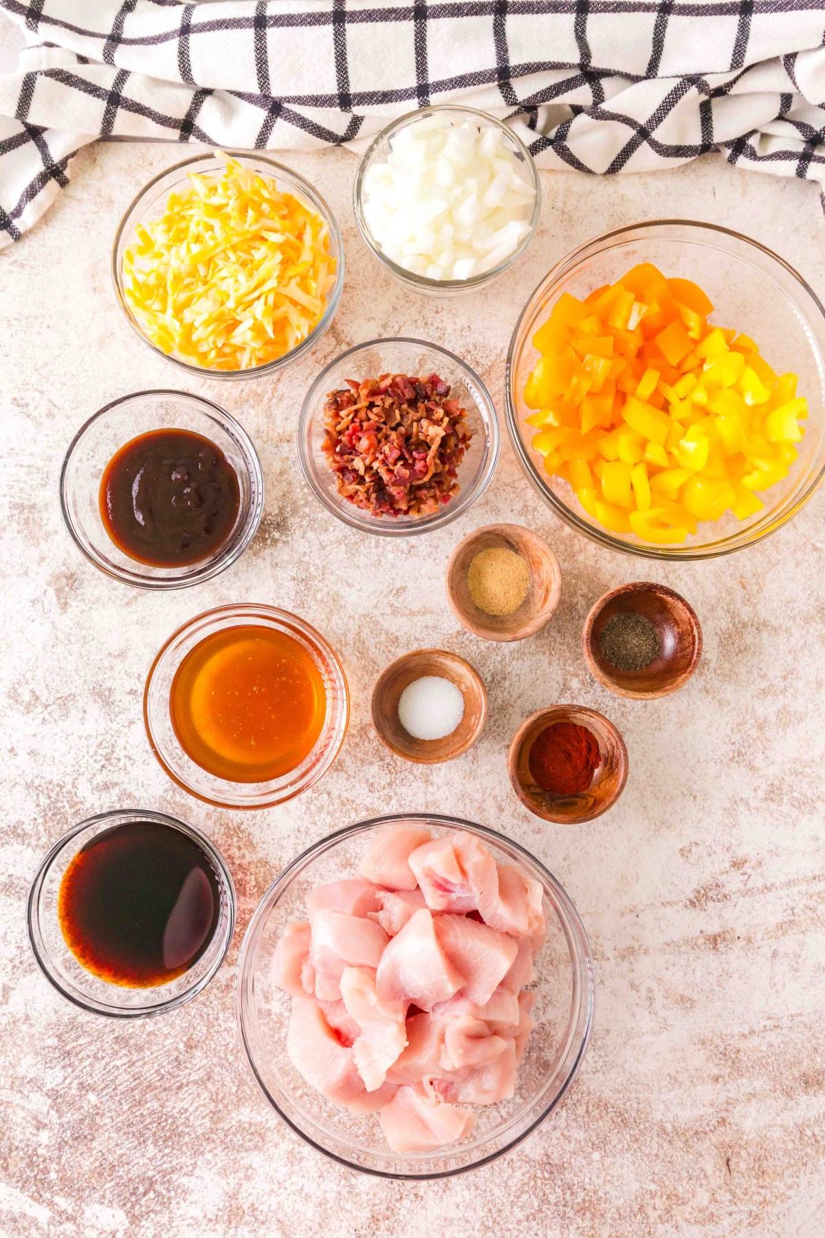 Bowls of chopped chicken, yellow peppers, cheese, onion, bacon, and various sauces and spices on a counter.