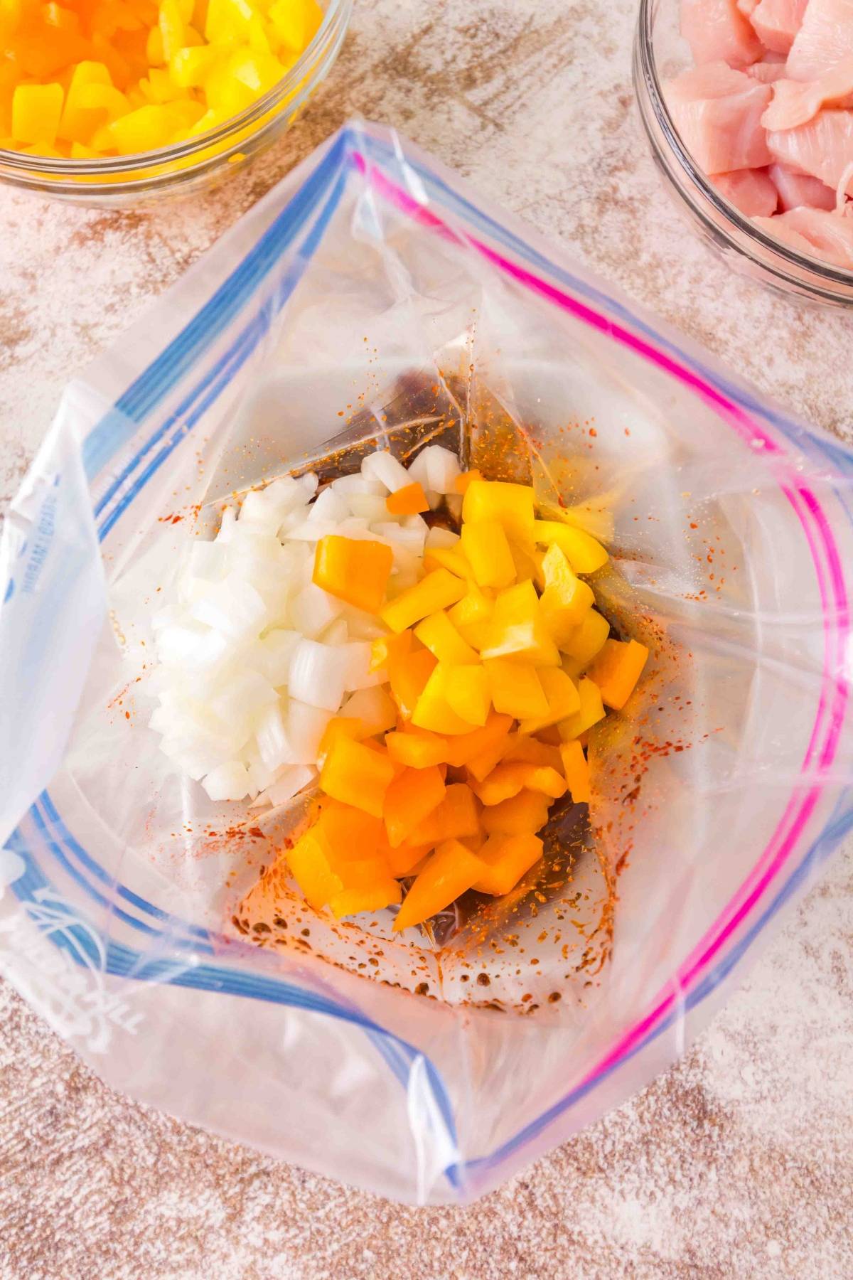 Chopped onions and yellow bell peppers in a zipper bag with seasonings, on a countertop with ingredients nearby.