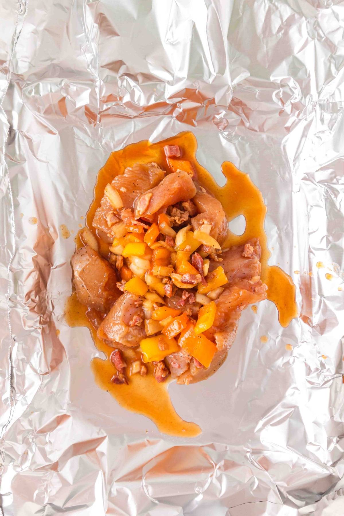 Raw marinated chicken pieces with chopped yellow peppers on aluminum foil, ready to be wrapped and cooked.
