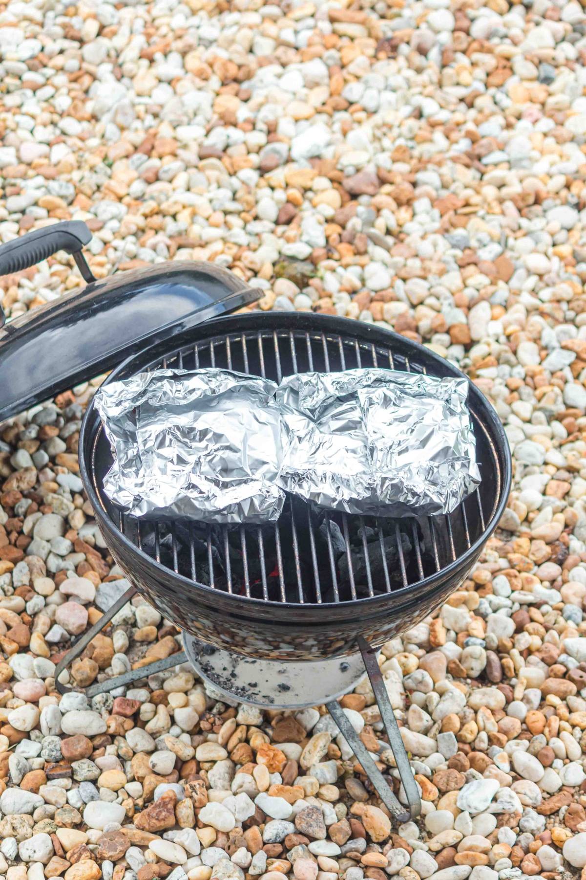 Two foil-wrapped packets cooking on a small charcoal grill with pebbles covering the ground around it.